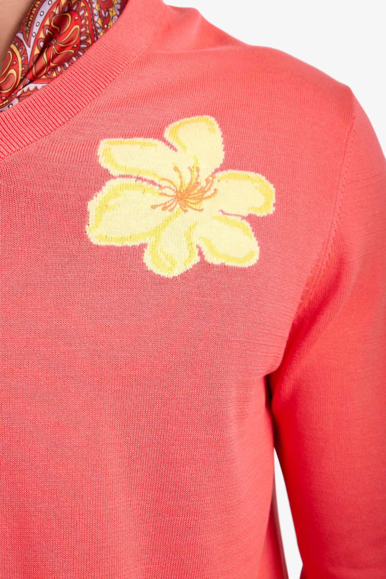 A close-up of the Divino V-Neck Sweater in coral pink, featuring a bold yellow flower on the chest, styled with a patterned red scarf.