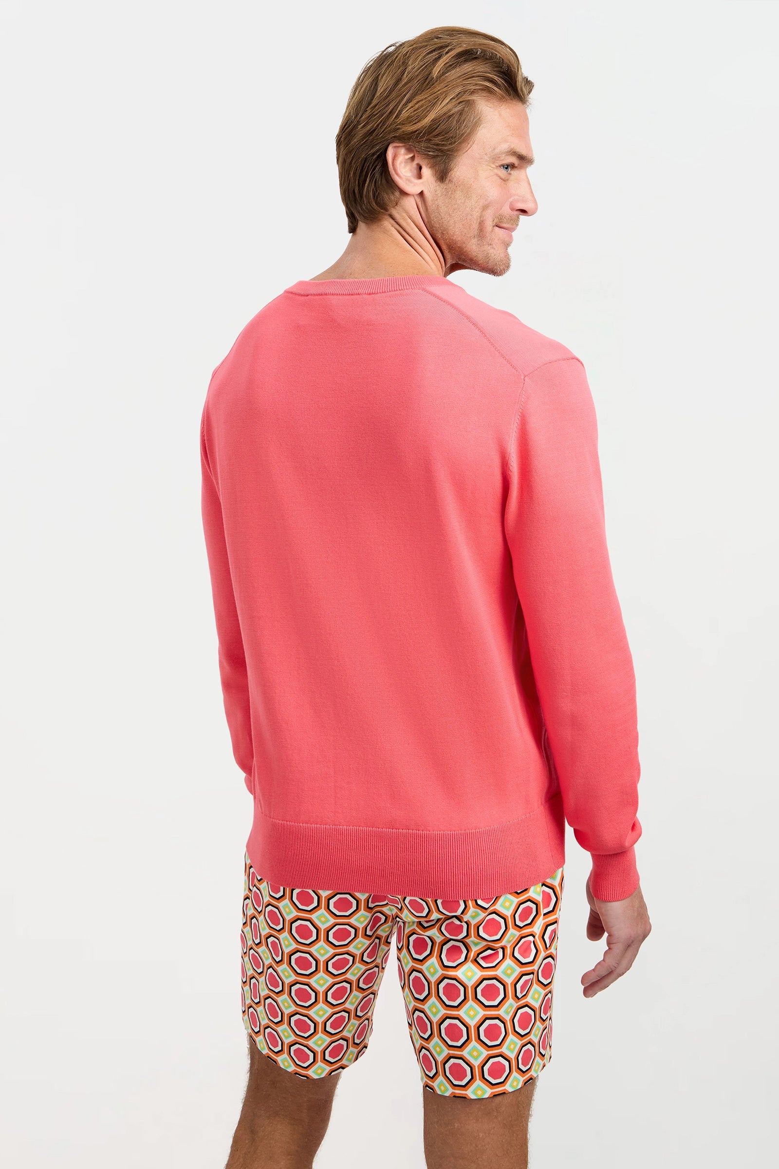A man stands facing away, wearing the Divino V-Neck Sweater in bright pink and colorful, patterned shorts with overlapping circles in red, yellow, and white against a plain white background.