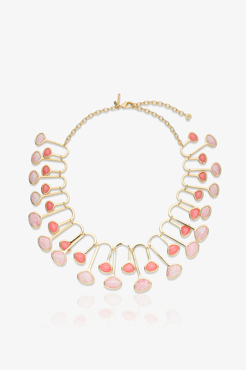 The Flamingo Necklace is a gold necklace with a chain clasp, featuring 22 pink and coral oval stones set in a fan-like pattern. The stones are arranged in pairs along curved gold frameworks, creating a bold, elegant statement piece.