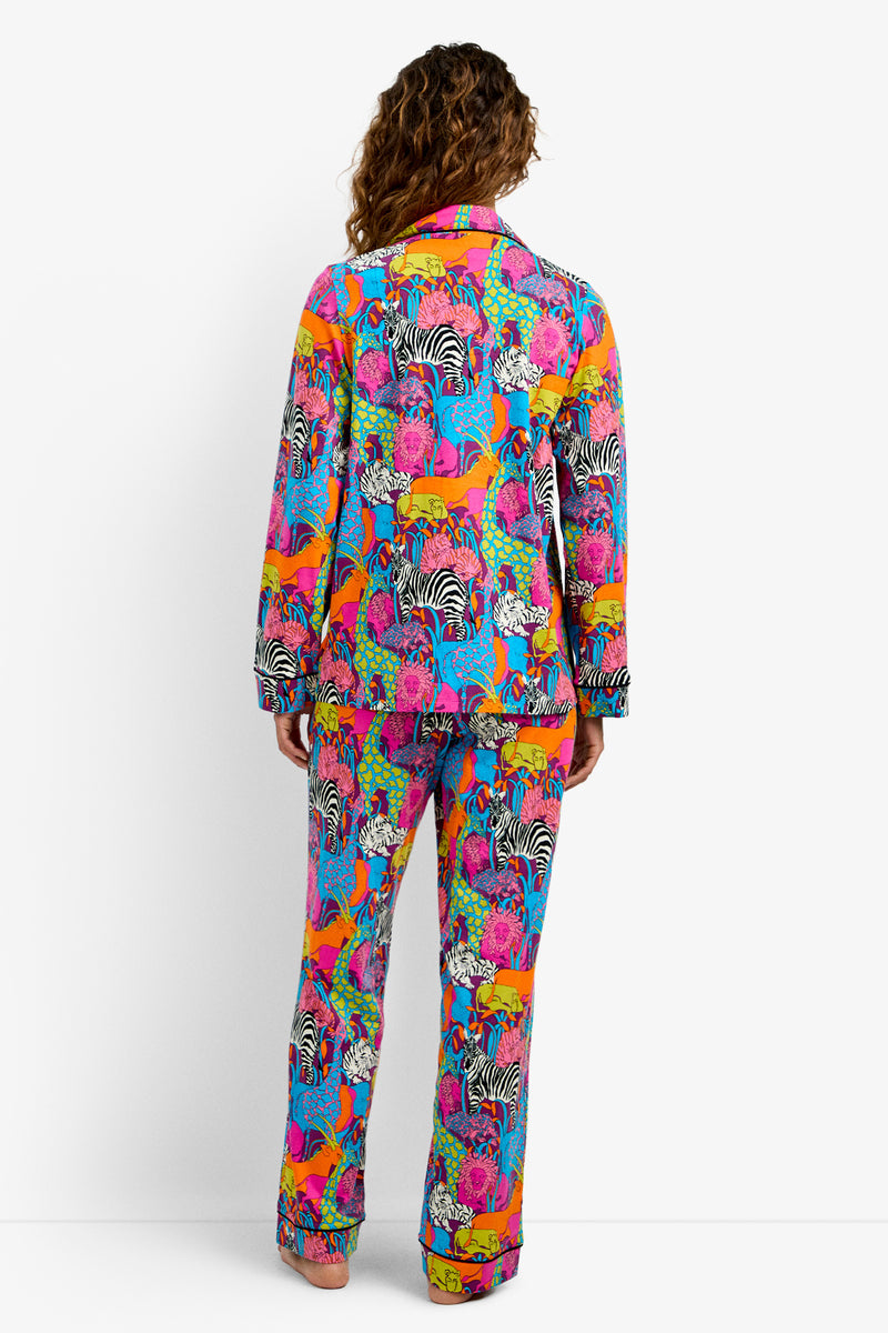Someone stands facing away, wearing the Classic PJ Set - Jungle, featuring a vibrant animal pattern with zebras, tigers, and more in pink, orange, blue, green, and yellow on a white background.
