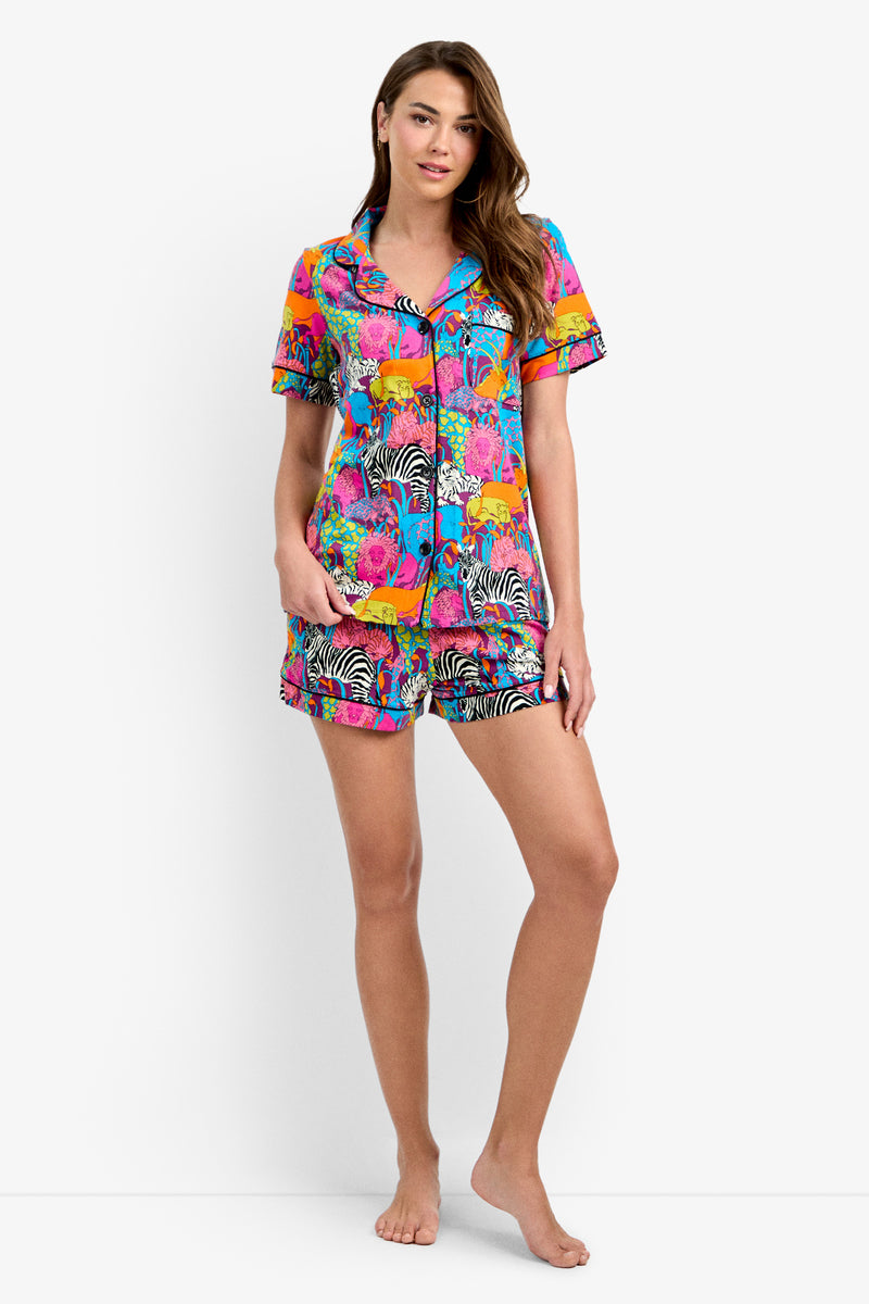 A woman stands barefoot against a white background wearing the Shorty PJ Set - Jungle, a colorful short-sleeve pajama set with a vibrant zebra and floral print.