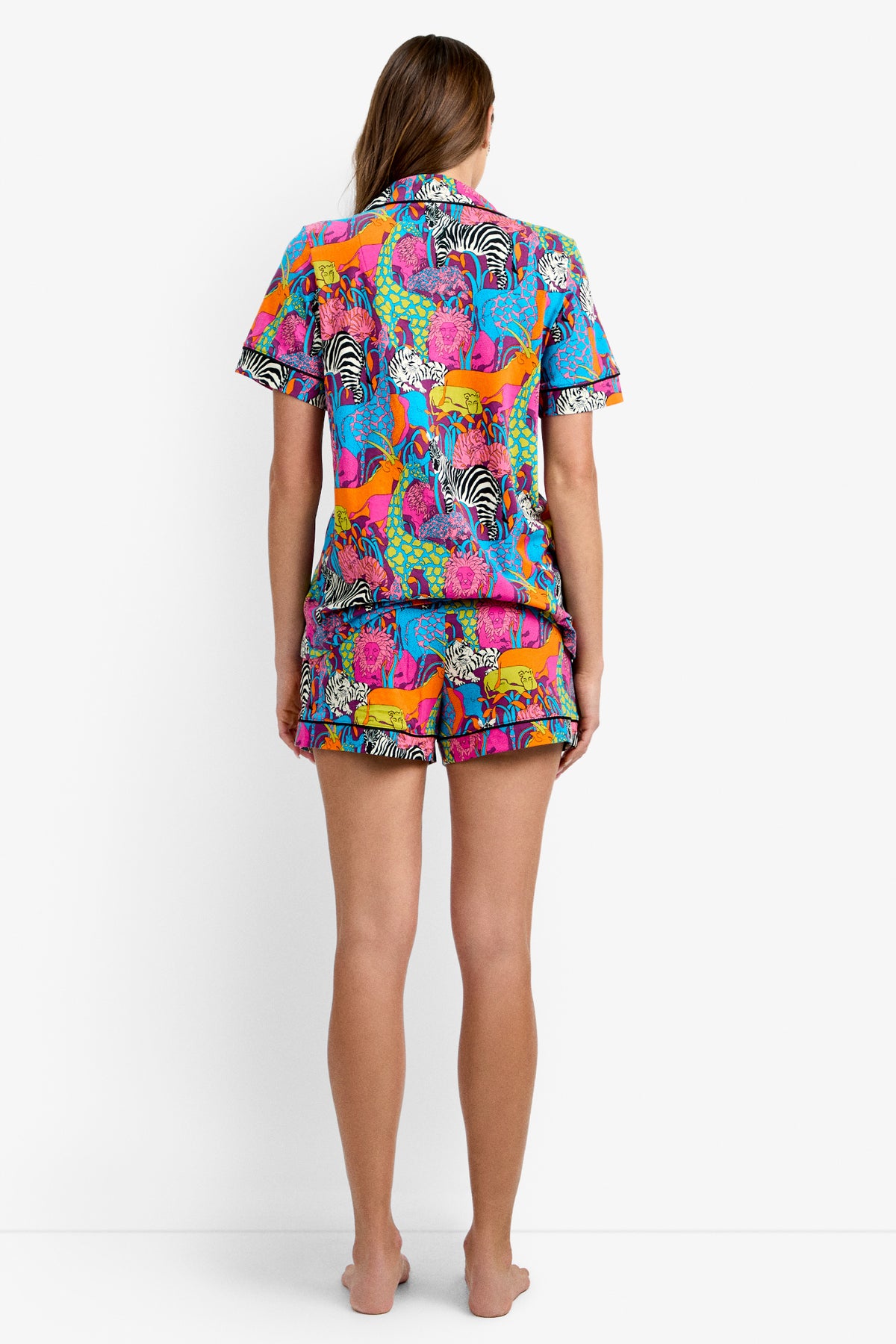 A person stands barefoot facing away, wearing the Shorty PJ Set - Jungle, a colorful short-sleeve pajama set featuring vibrant animal prints like zebras and big cats. The background is plain white.