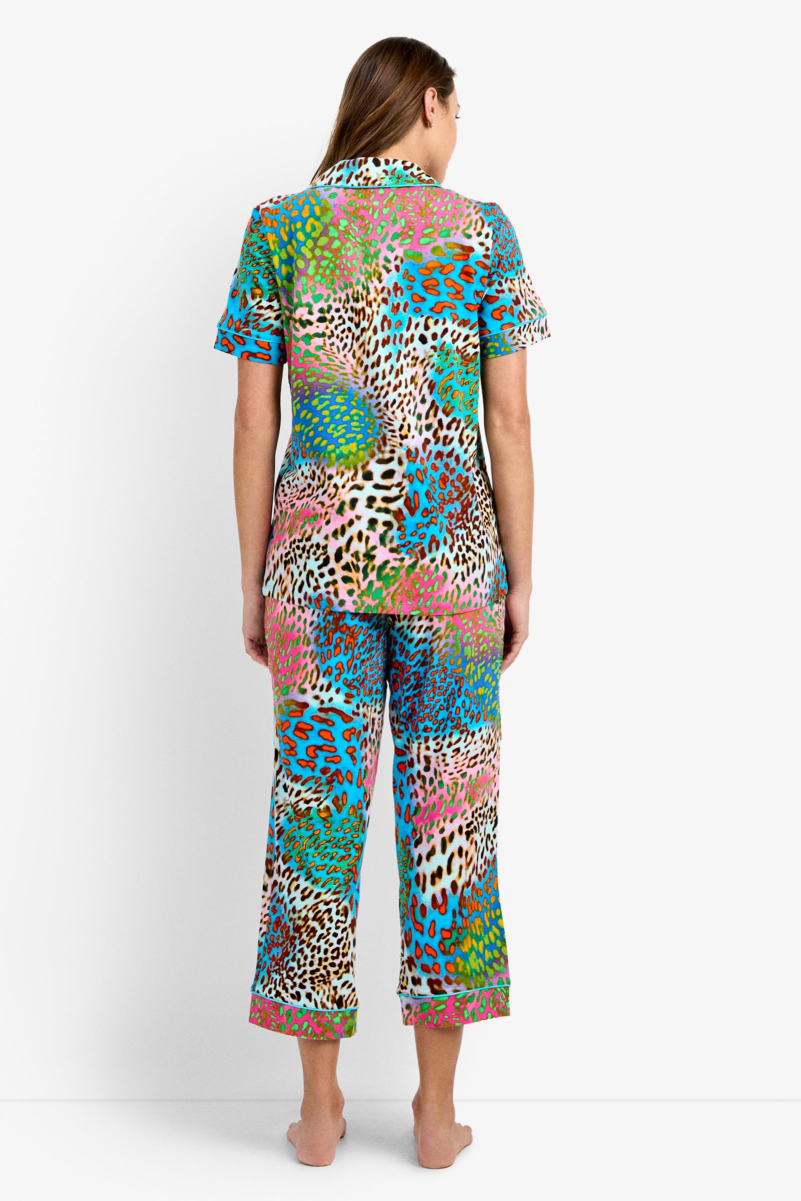 A woman stands barefoot facing away, wearing the Crop Classic PJ Set - Spray Leopard, a colorful short-sleeve pajama set featuring a bold multicolored leopard print on both the top and cropped pants.