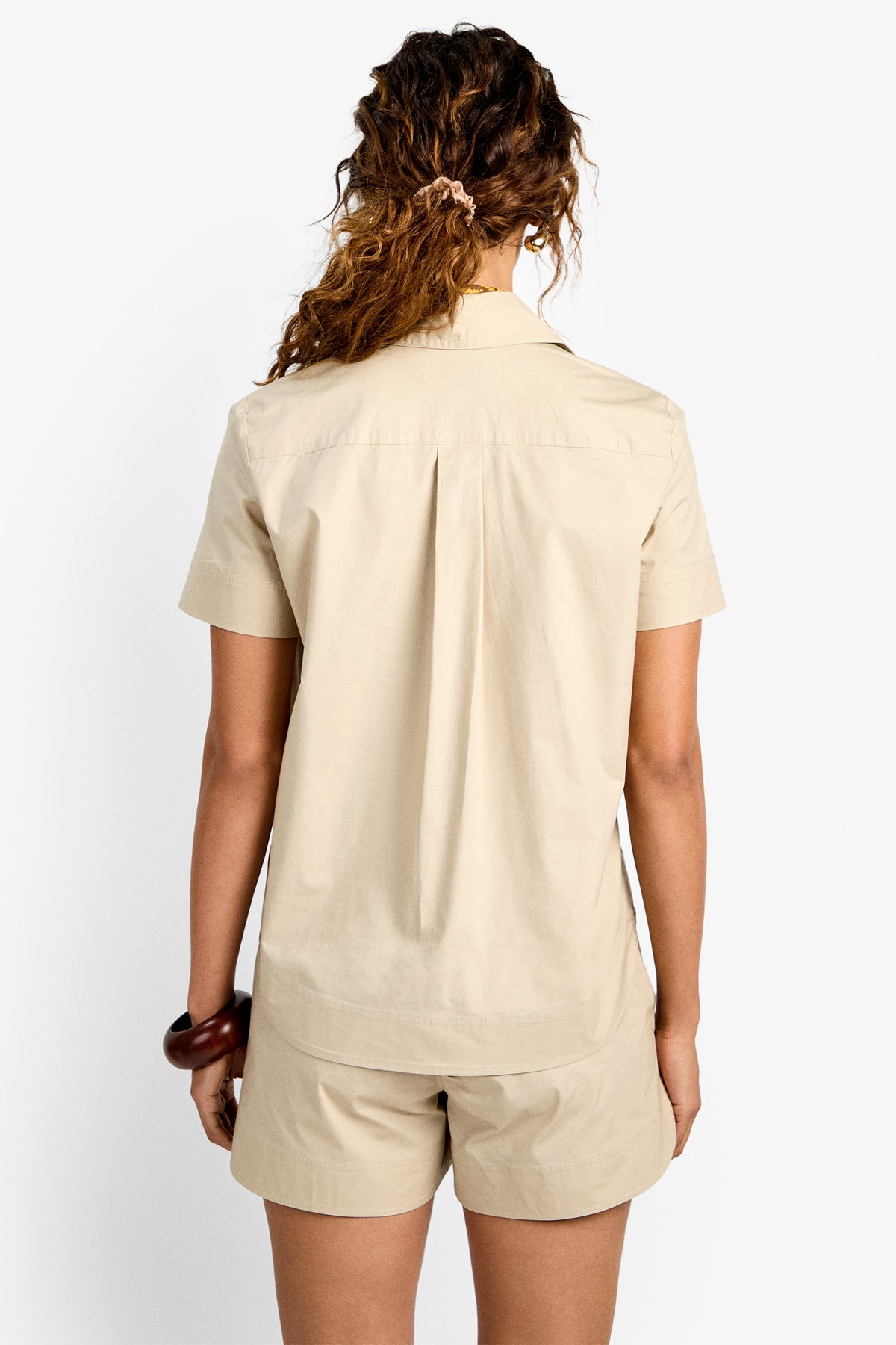 A person with wavy hair in a loose ponytail is seen from the back, wearing the Coral Top, a short-sleeve button-up shirt, and matching shorts in beige against a plain white background.