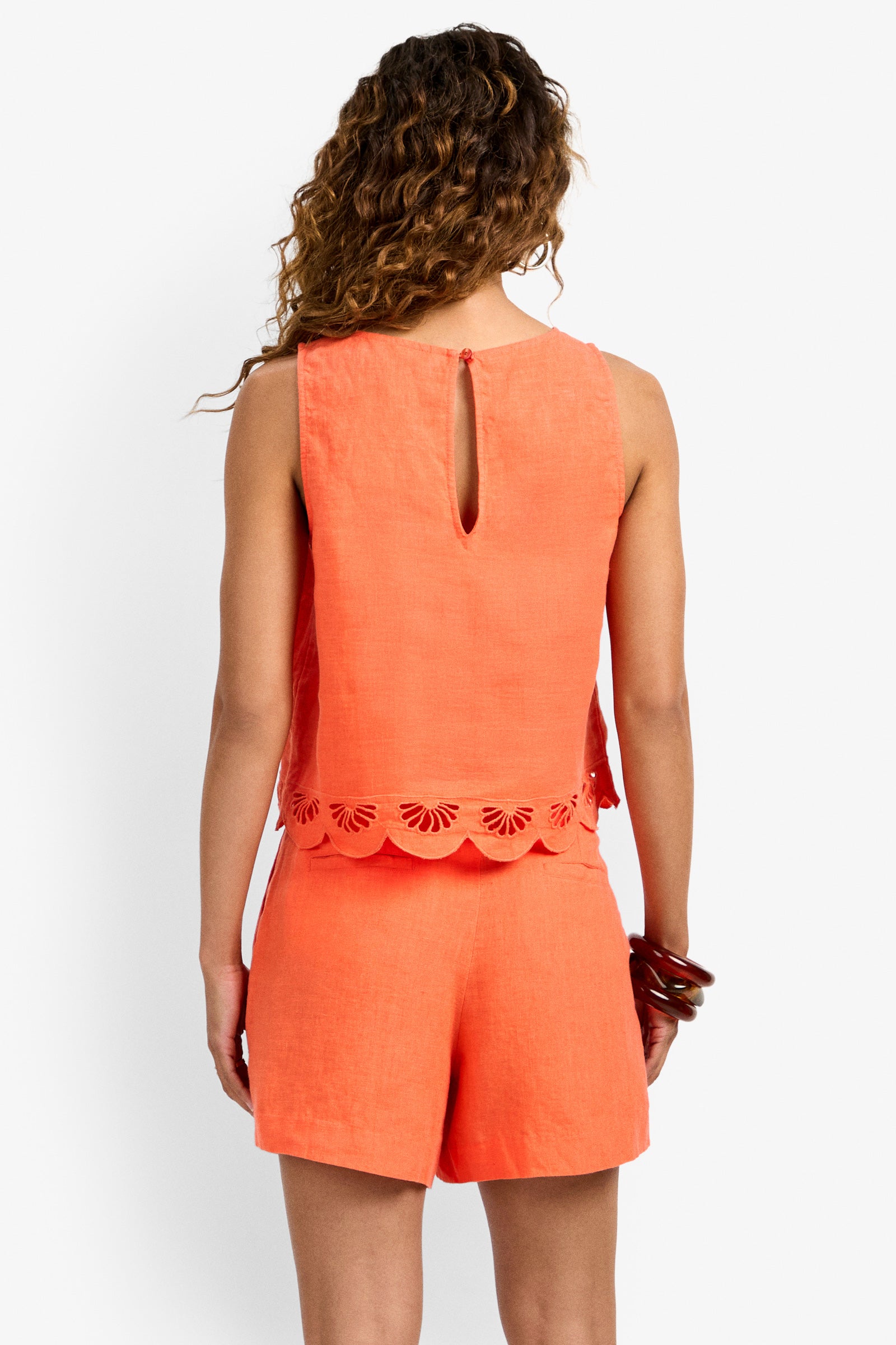 A woman with wavy brown hair is seen from the back wearing the sleeveless coral Kailani Top with a scalloped, embroidered hem and matching shorts. She holds brown sunglasses in her right hand.