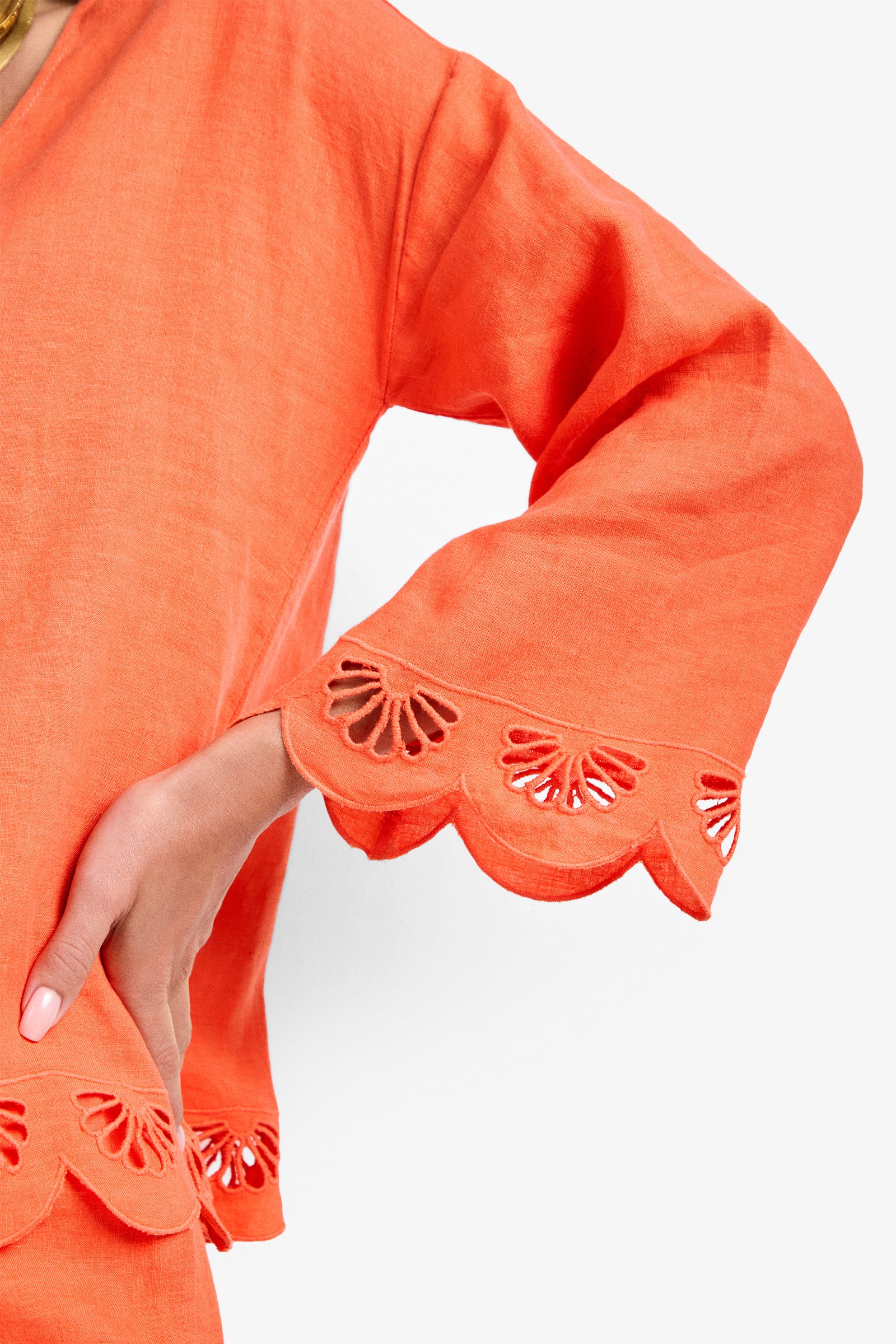 A close-up of a person wearing the Mar Top, an orange long-sleeve shirt with scalloped and cutout floral patterns on the sleeve cuffs, their hand resting on their hip.
