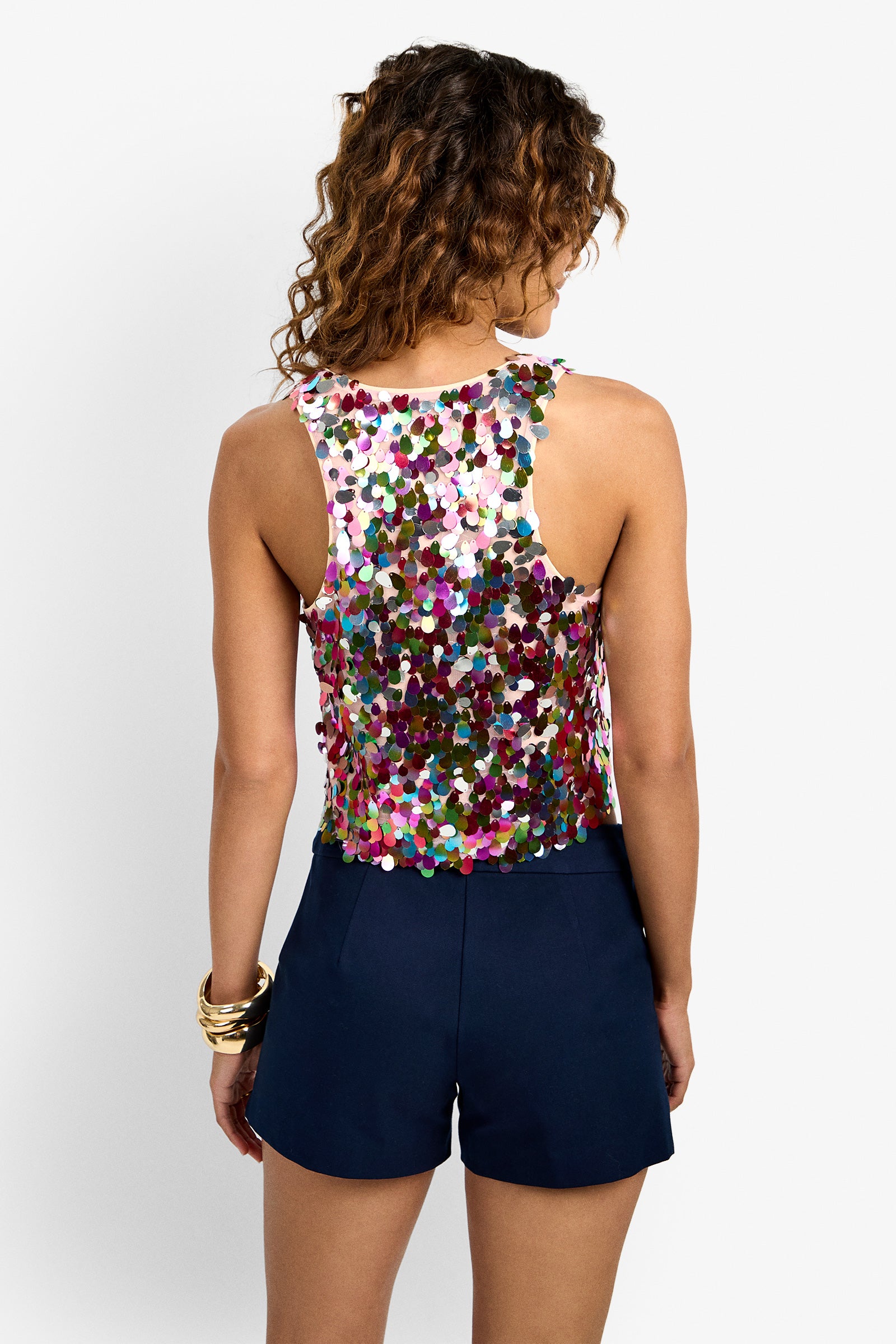 A woman with curly hair faces away, wearing the colorful Nerissa Top and dark navy shorts. She accessorizes with gold bangles and stands against a plain white background.