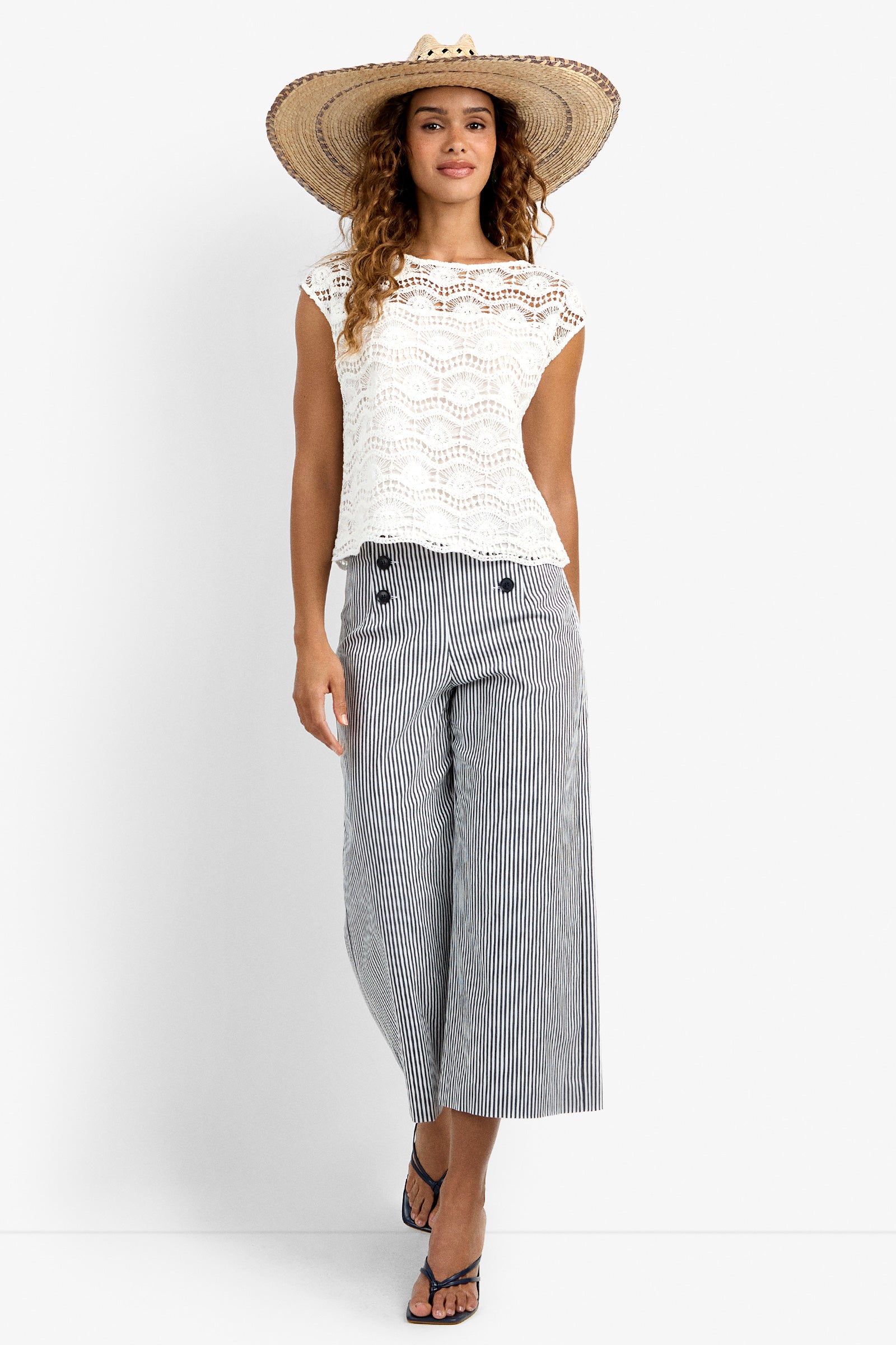 A woman wearing the Neptune Top, a large straw hat, wide-leg striped pants, and flip-flops stands against a plain background, smiling gently.
