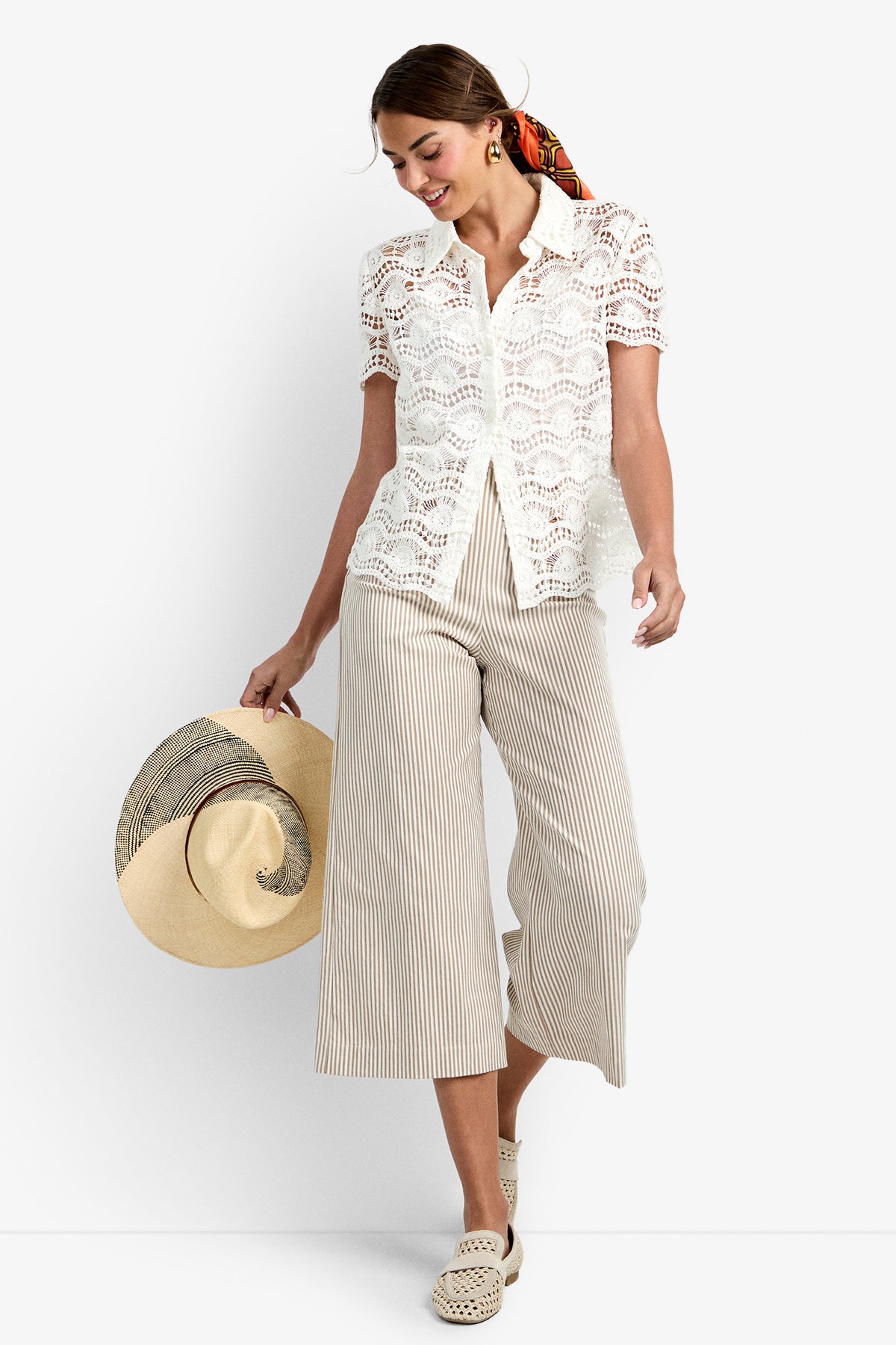 Wearing the Triton Top in WHITEWASH, a woman paired it with beige and white striped wide-leg cropped pants, woven light shoes, and a colorful scarf in her hair as she smiles and looks down, holding a large floppy hat.