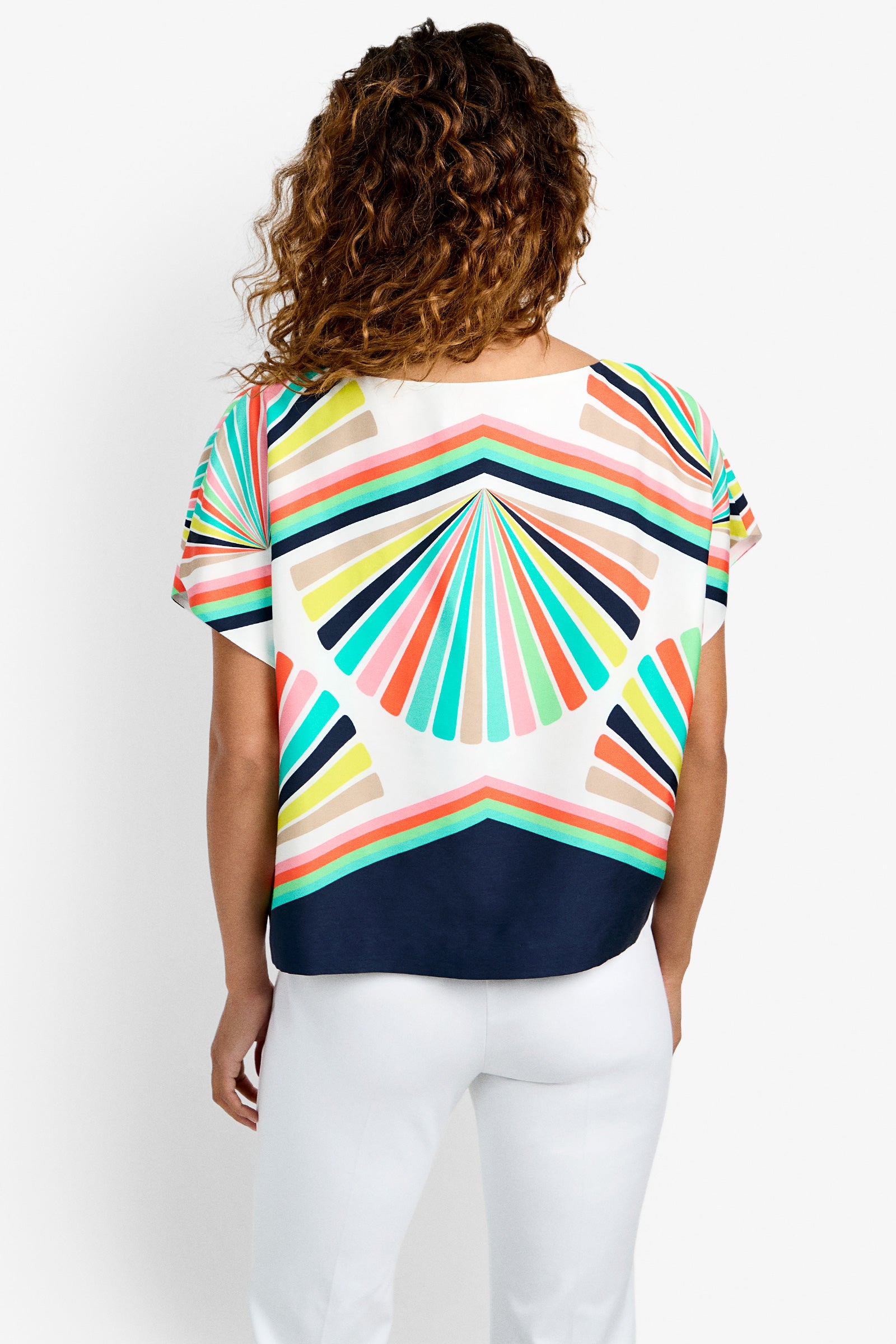 Seen from behind against a white background, a person with curly hair wears the Breezy Top—featuring colorful geometric fan-shaped patterns in aqua, coral, yellow, and navy—paired with white pants.