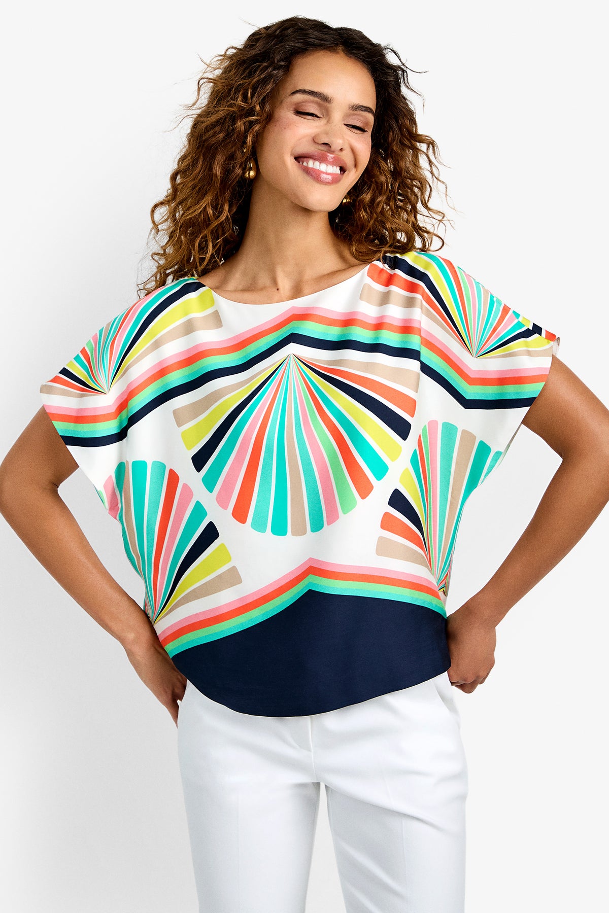 A smiling woman with curly hair models the Breezy Top, a colorful blouse with geometric patterns and wide sleeves, paired with white pants. She stands confidently with hands on hips against a plain white background.