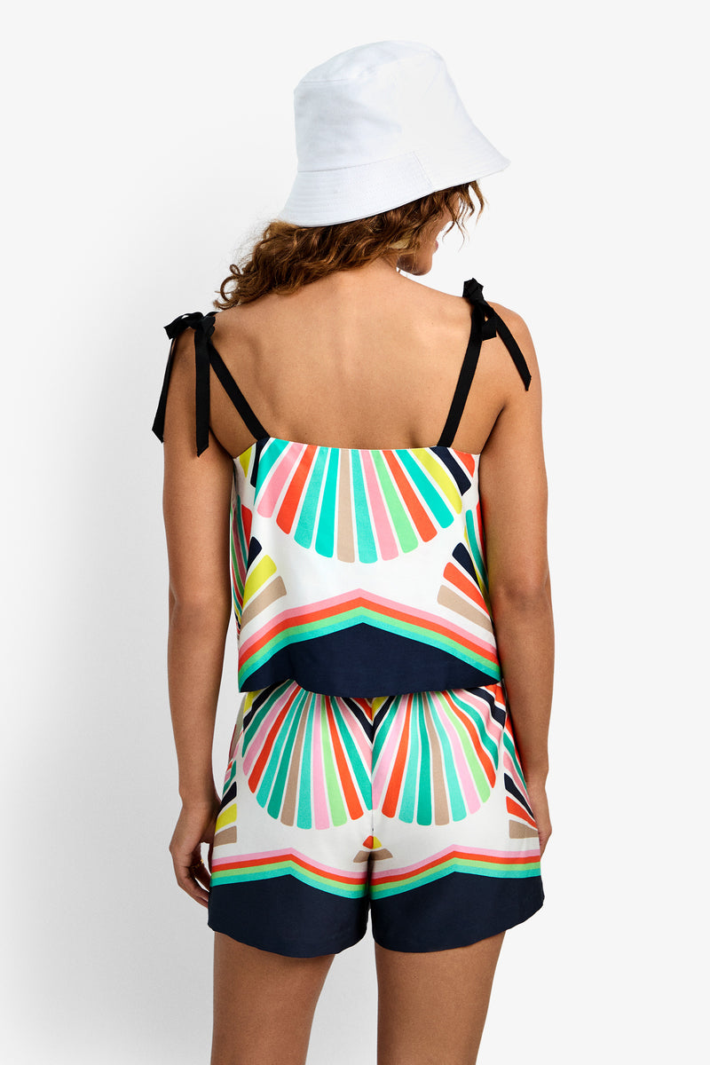A woman faces away wearing a white bucket hat and the Marella Top—a colorful, geometric-patterned tank with black shoulder ties, multicolored stripes, and navy blue accents—paired with matching shorts.