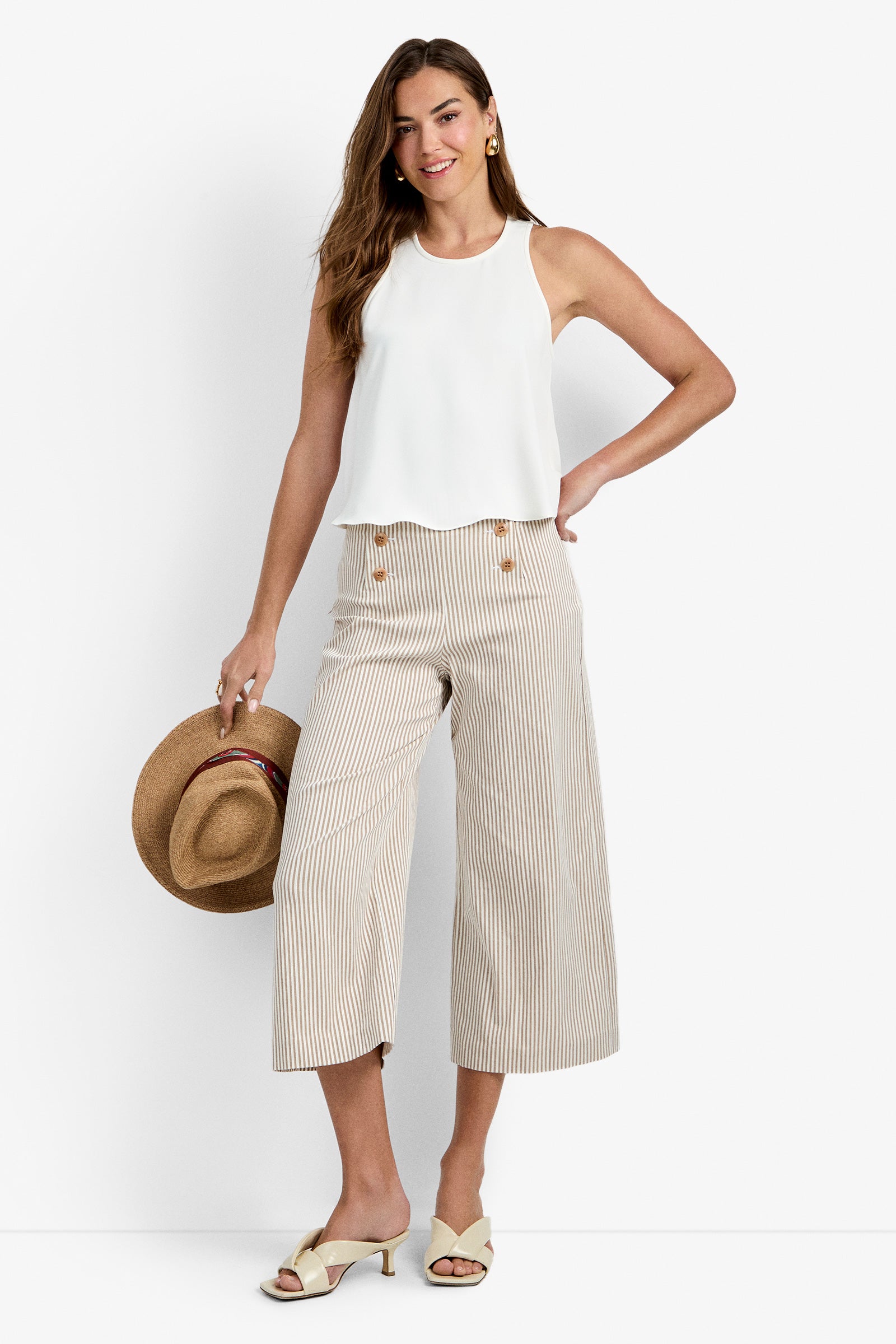 A woman smiles in a sleeveless white top and the Saltie Pant—wide-leg beige striped pants. She holds a straw hat, wears beige sandals, has long hair, and accessorizes with gold earrings.