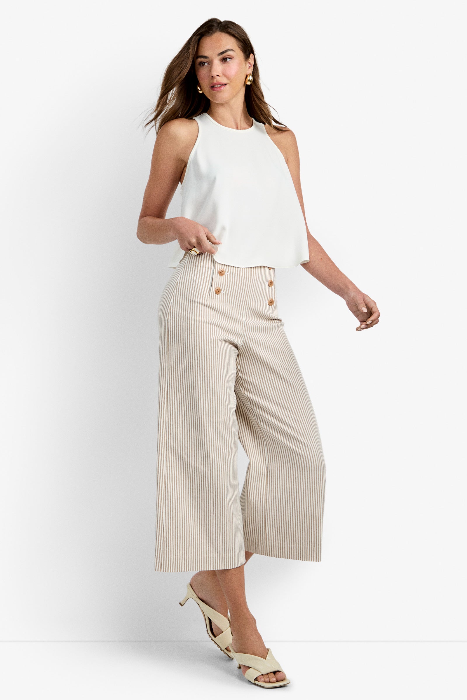 A woman poses in the Zuria Top in Whitewash, paired with high-waisted, wide-leg beige striped pants with buttons. She completes the look with cream heels, gold earrings, and softly styled hair worn down.