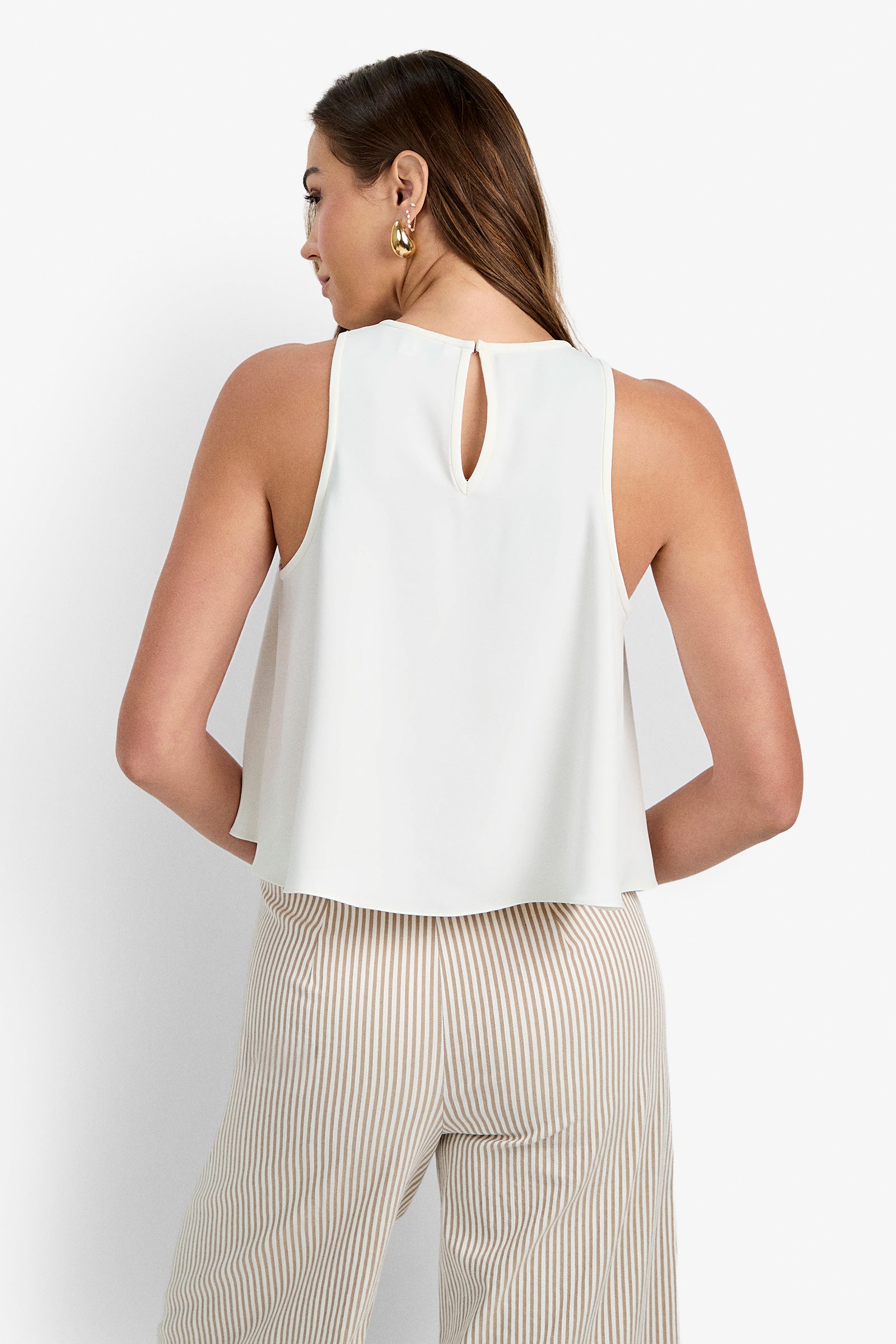 A woman with long brown hair and gold hoop earrings is shown from the back wearing the sleeveless Zuria Top, featuring a white color and keyhole neckline, paired with beige and white vertical striped pants.
