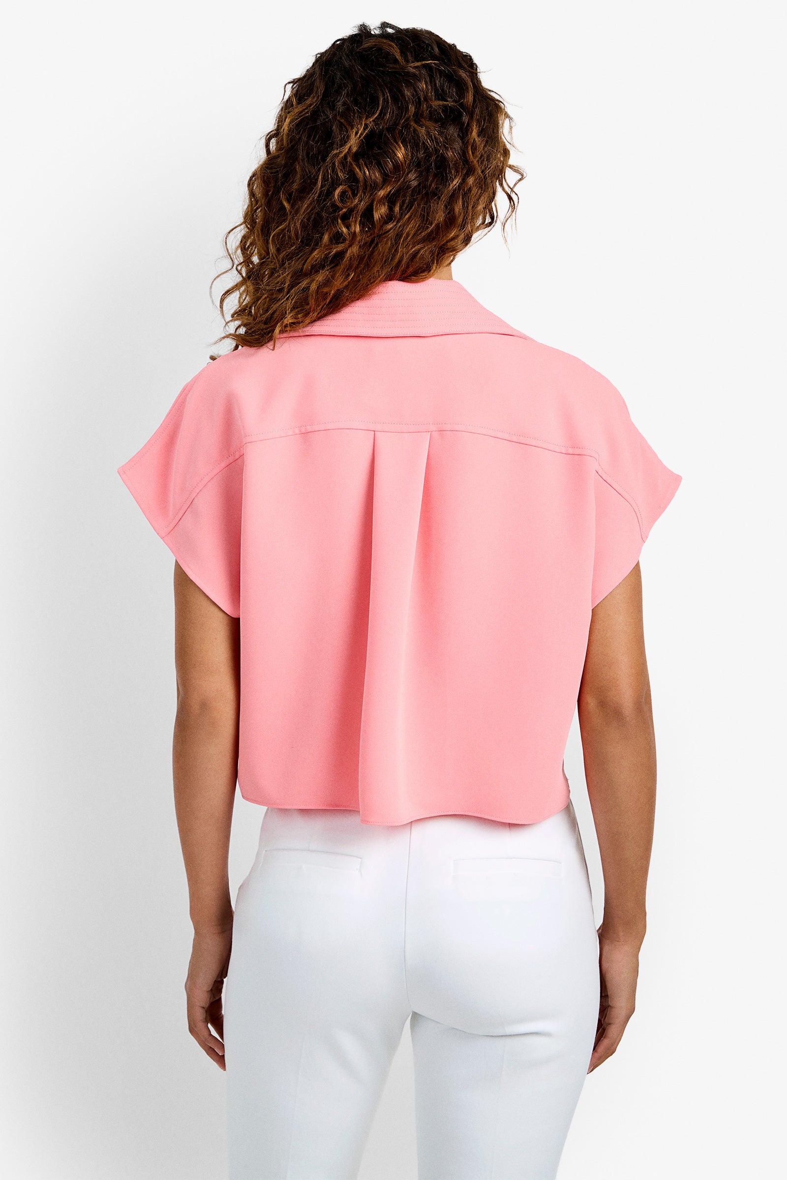 A person with curly brown hair is seen from behind, wearing the Stream Top—a short-sleeve, loose-fitting pink blouse—paired with white pants against a plain white background.