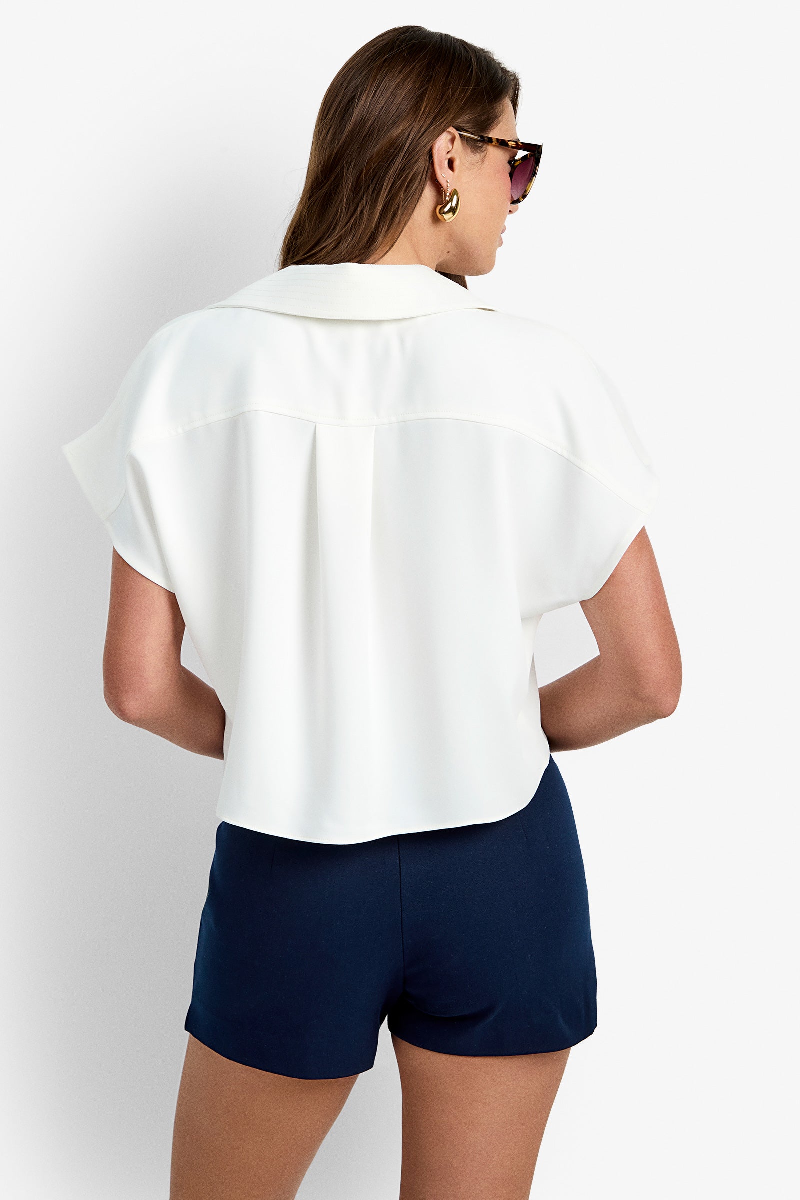 A woman shown from the back against a plain white background wears sunglasses, navy blue shorts, and the Stream Top, a white short-sleeve blouse.