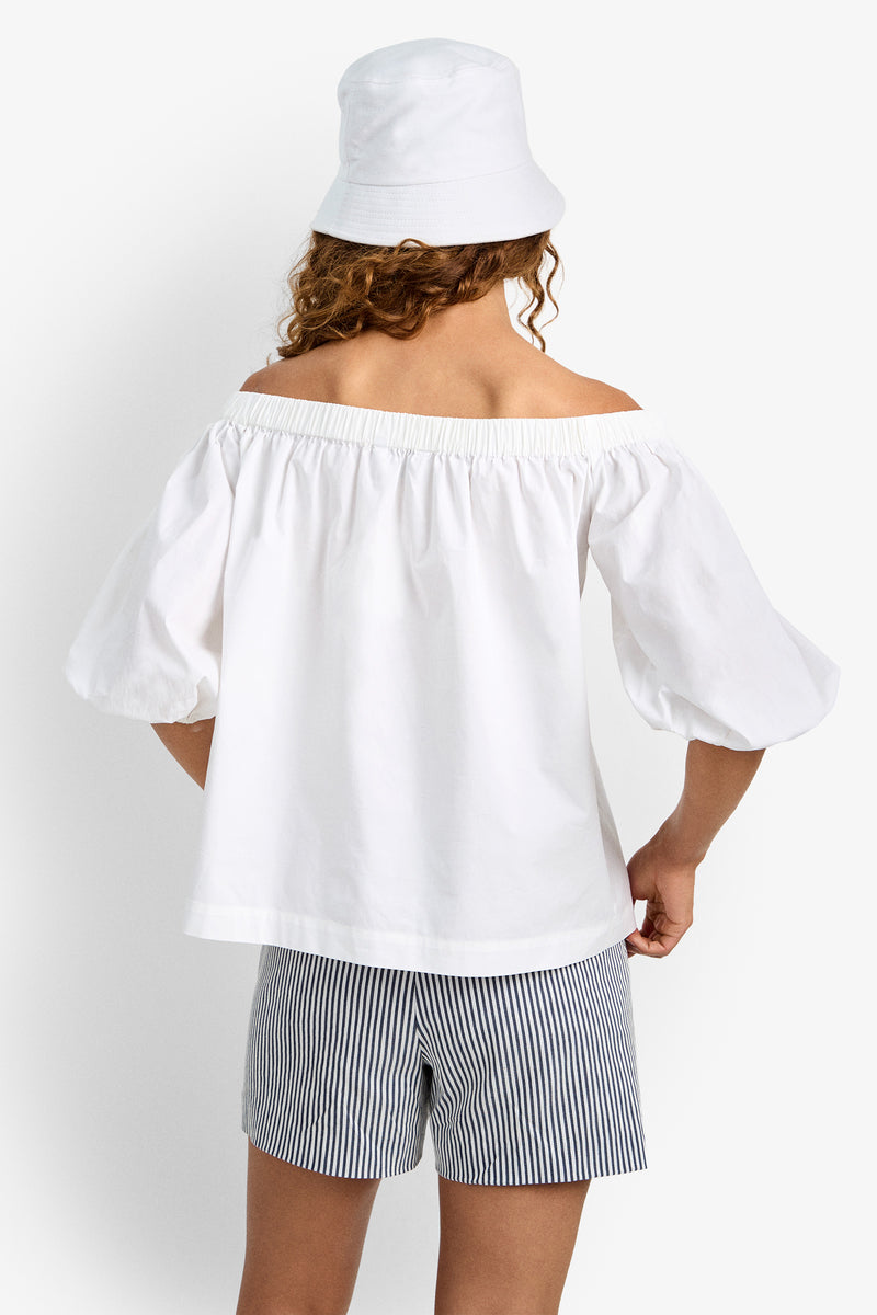 A person with curly hair, shown from behind against a plain background, is wearing the white off-the-shoulder Cove Top, a white bucket hat, and blue-and-white striped shorts.