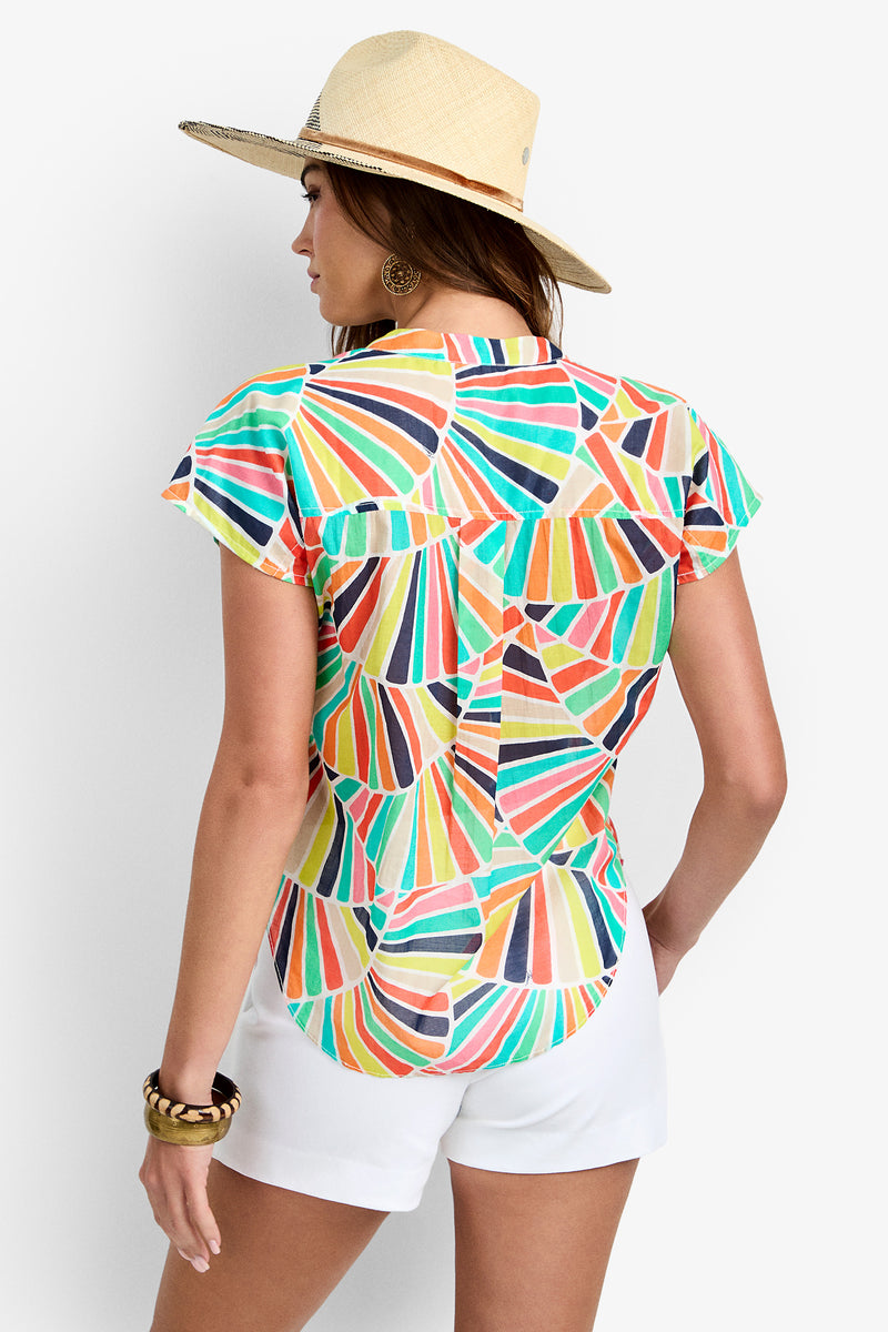 A woman stands facing away against a plain white background, wearing the Krill Top—a colorful geometric-patterned, short-sleeved blouse—paired with a wide-brimmed straw hat and white shorts.