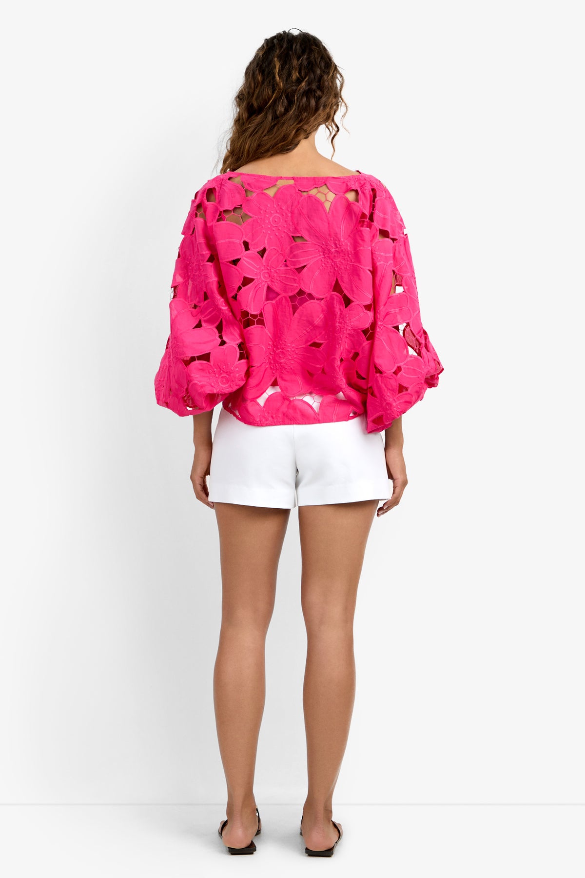 A woman with wavy, shoulder-length hair stands facing away, wearing the bright pink Coastal Top with floral cutouts, puffy sleeves, and white shorts. She completes the look with flat sandals against a plain white background.