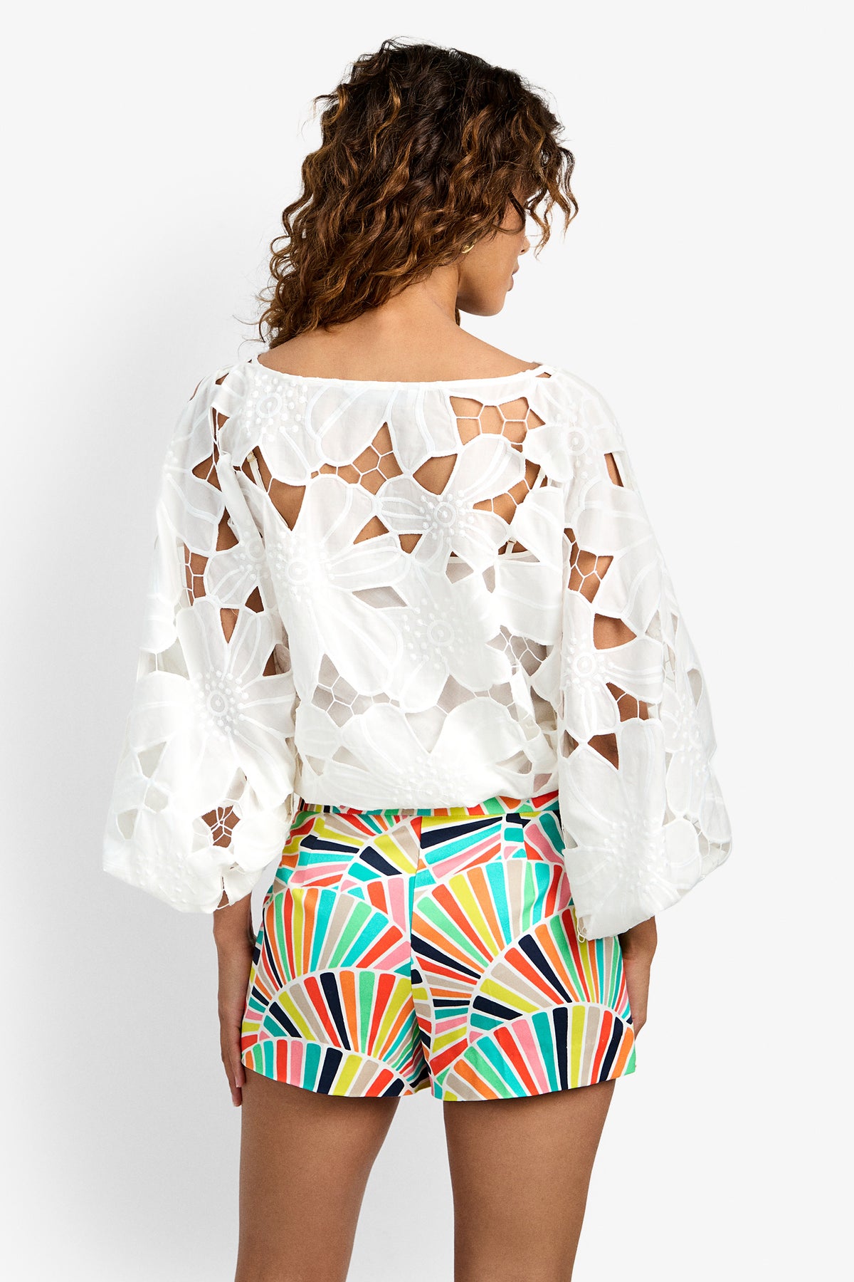 A woman with curly hair stands facing away, wearing the white Coastal Top and colorful, geometric-patterned shorts against a plain white background.