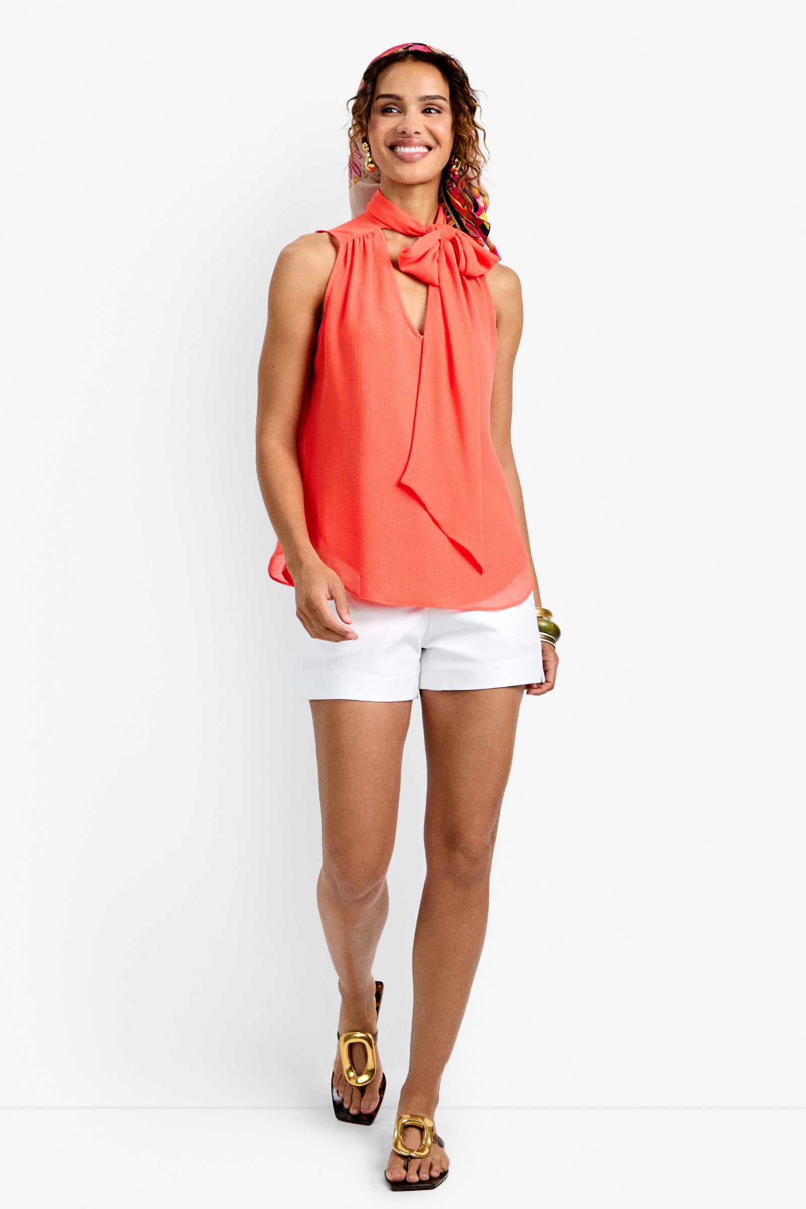 A smiling woman wears the Alora Top in CALYPSO CORAL, paired with white shorts, gold sandals, a headband, and bracelets, standing against a plain white background.
