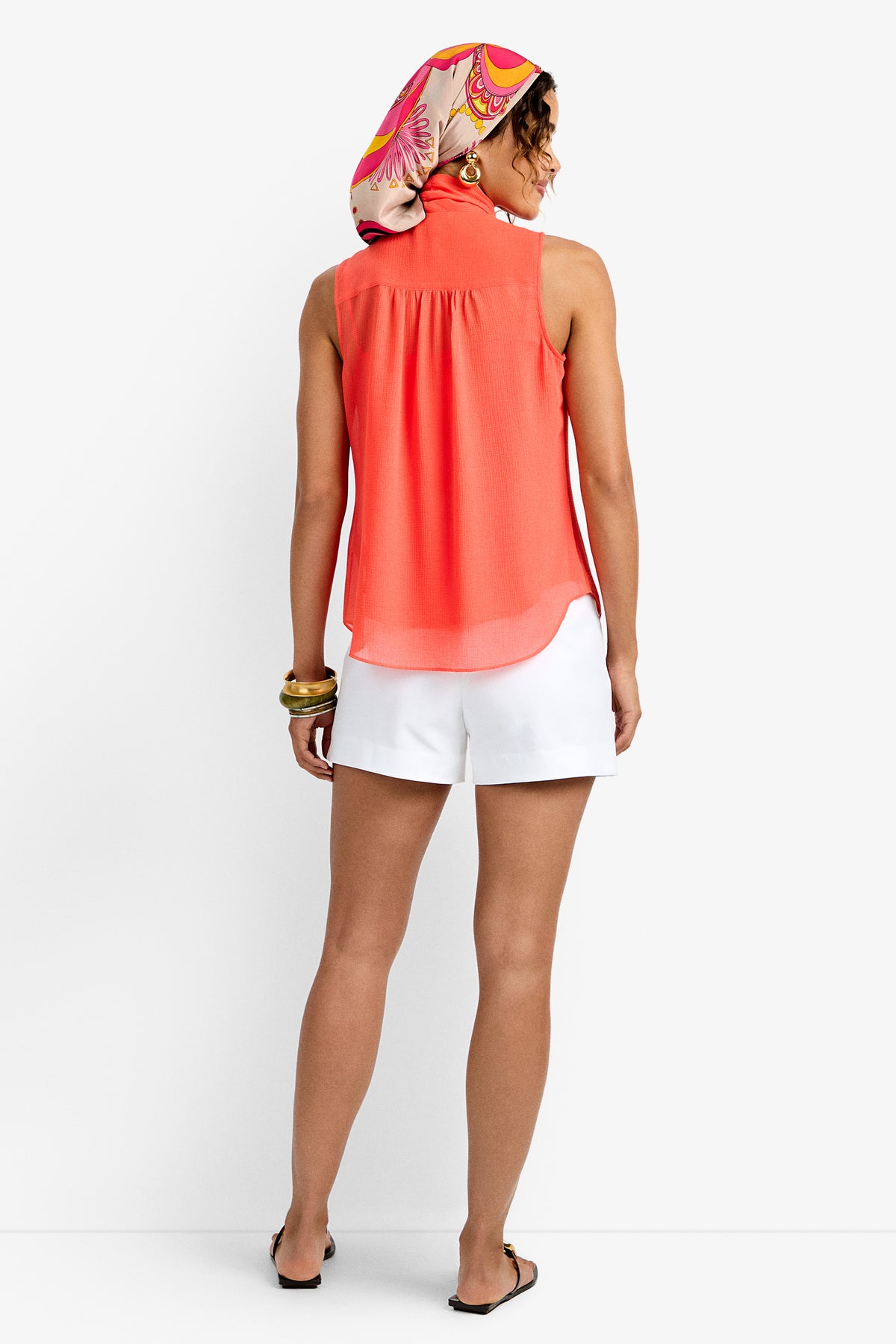 A woman faces away in the Alora Top, a sleeveless coral blouse, paired with white shorts, sandals, gold bangles, and a colorful headscarf. The background is plain white.
