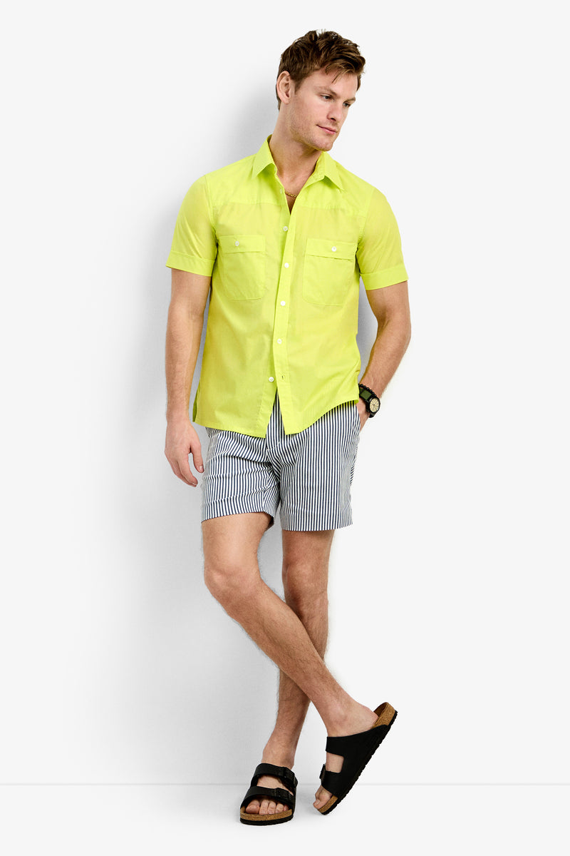 A man stands against a white background wearing the Will Shirt—a bright yellow short-sleeve button-up—paired with black-and-white striped shorts, black sandals, and a watch, with one hand in his pocket and legs crossed.