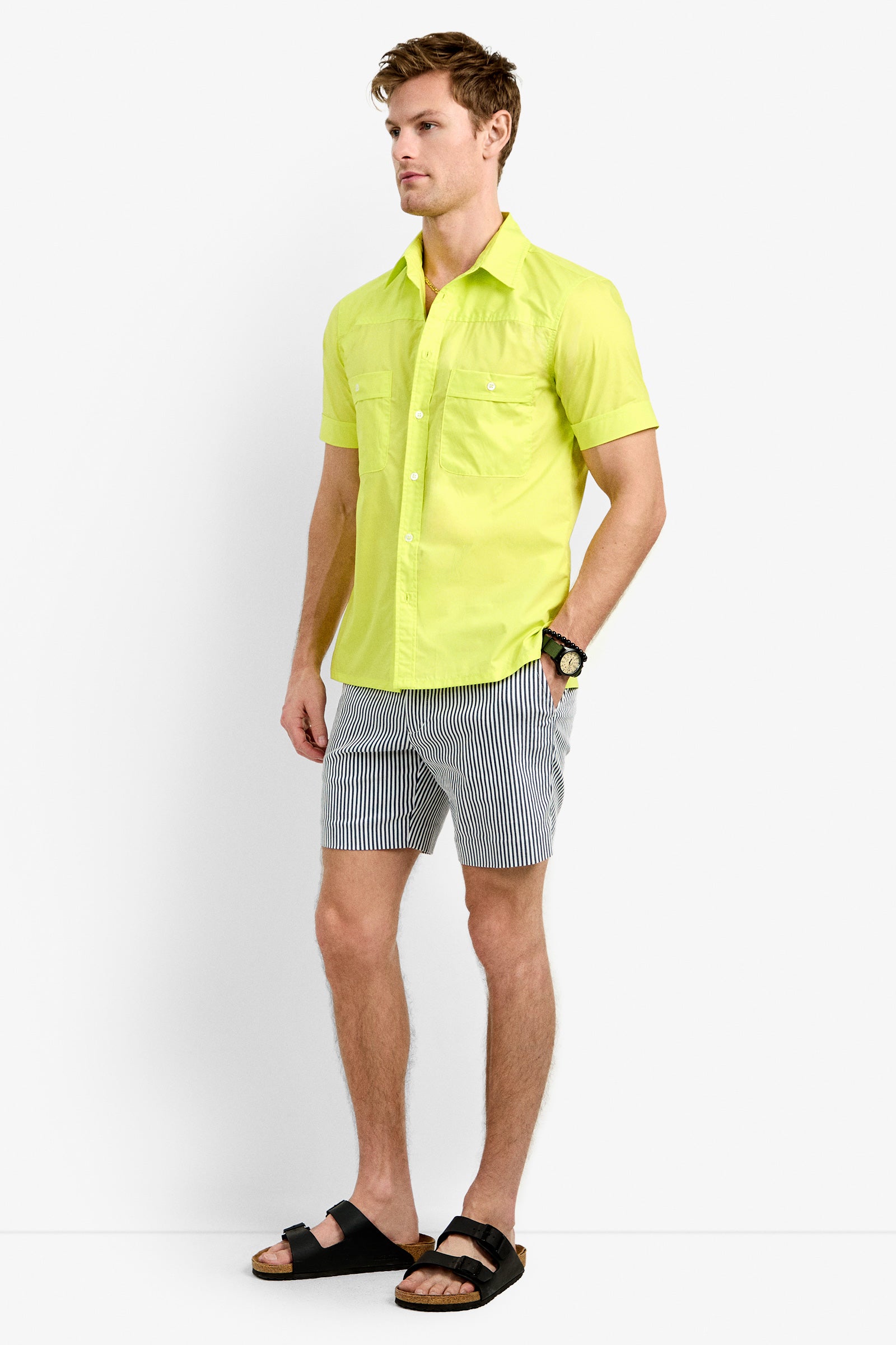 A man in the Will Shirt, a bright yellow short-sleeve button-up, pairs it with black-and-white striped shorts, black sandals, and a watch while standing against a plain white background.