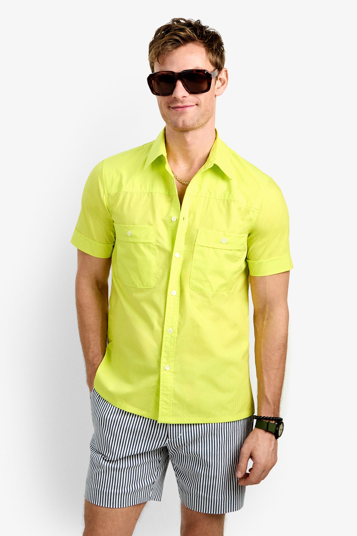 A man stands against a plain white background, smiling with his hands in his pockets, wearing the bright yellow Will Shirt, striped shorts, dark sunglasses, and a watch.
