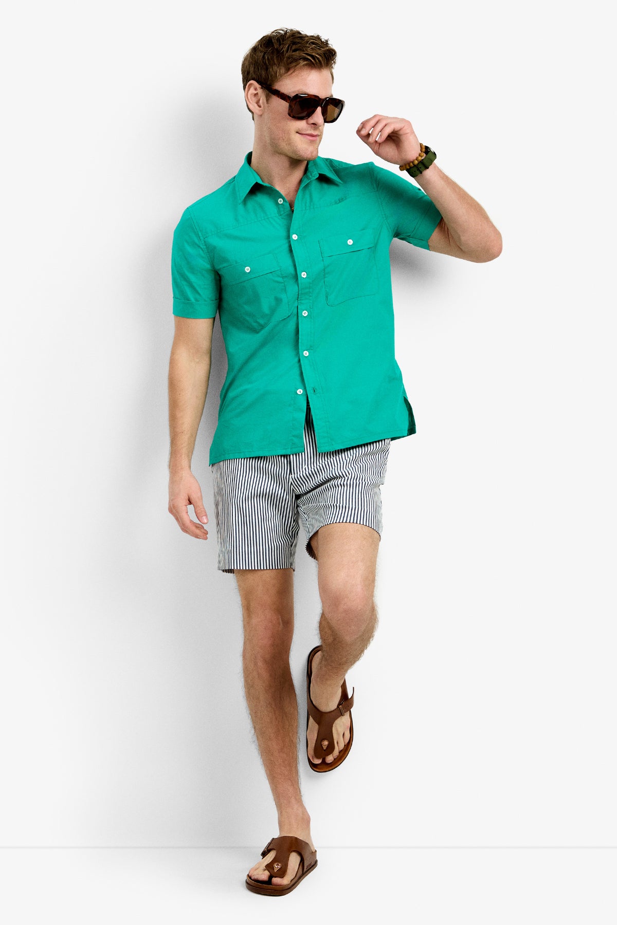 A man in the Will Shirt, a teal short-sleeve button-up, pairs it with striped shorts, brown sandals, and sunglasses. He stands against a white background looking relaxed and stylish in casual summer attire.