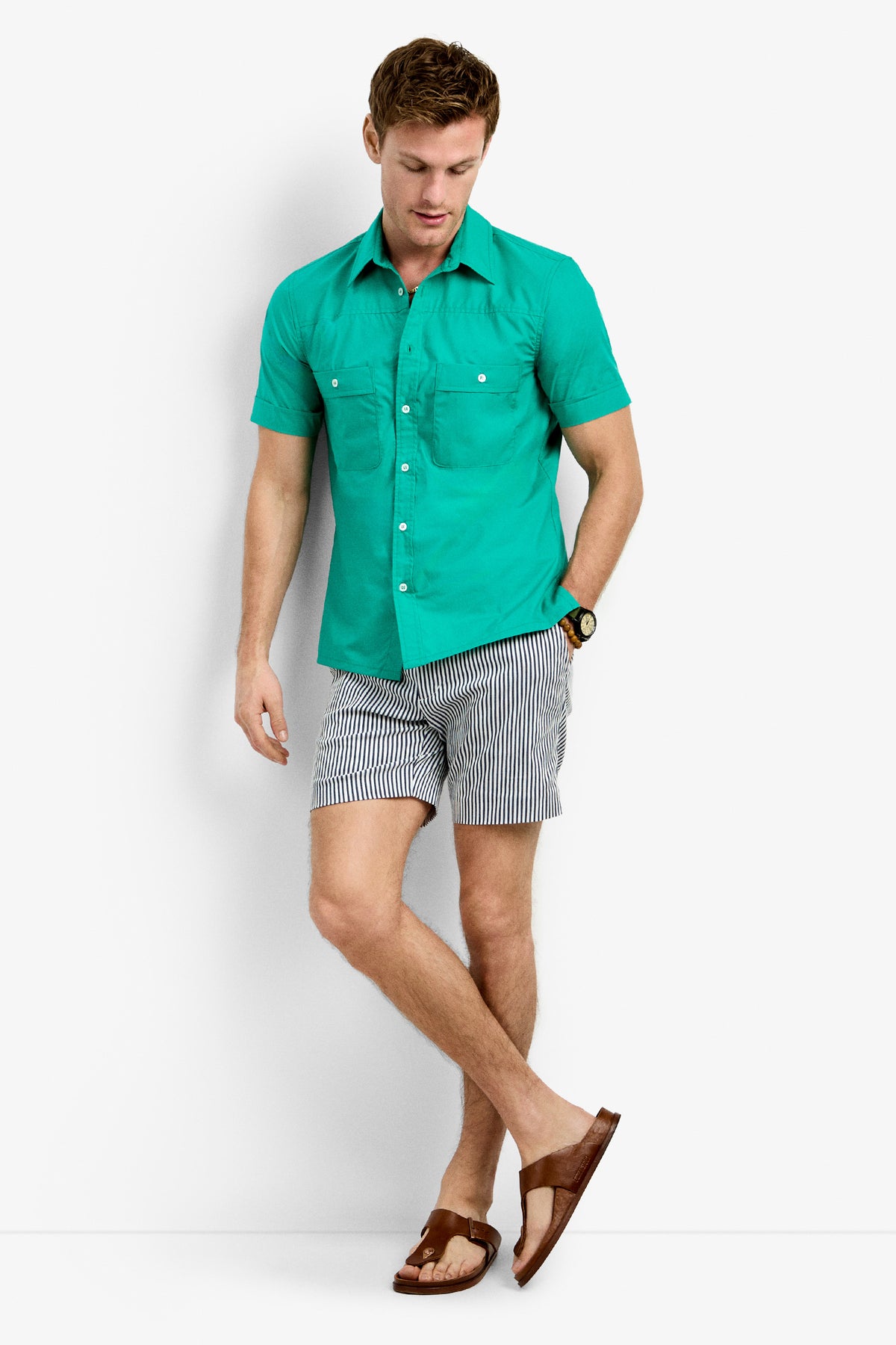 A man in the Will Shirt, a teal short-sleeve button-up, pairs it with black and white striped shorts, brown sandals, and a watch as he stands against a white background, looking down with one leg crossed over the other.