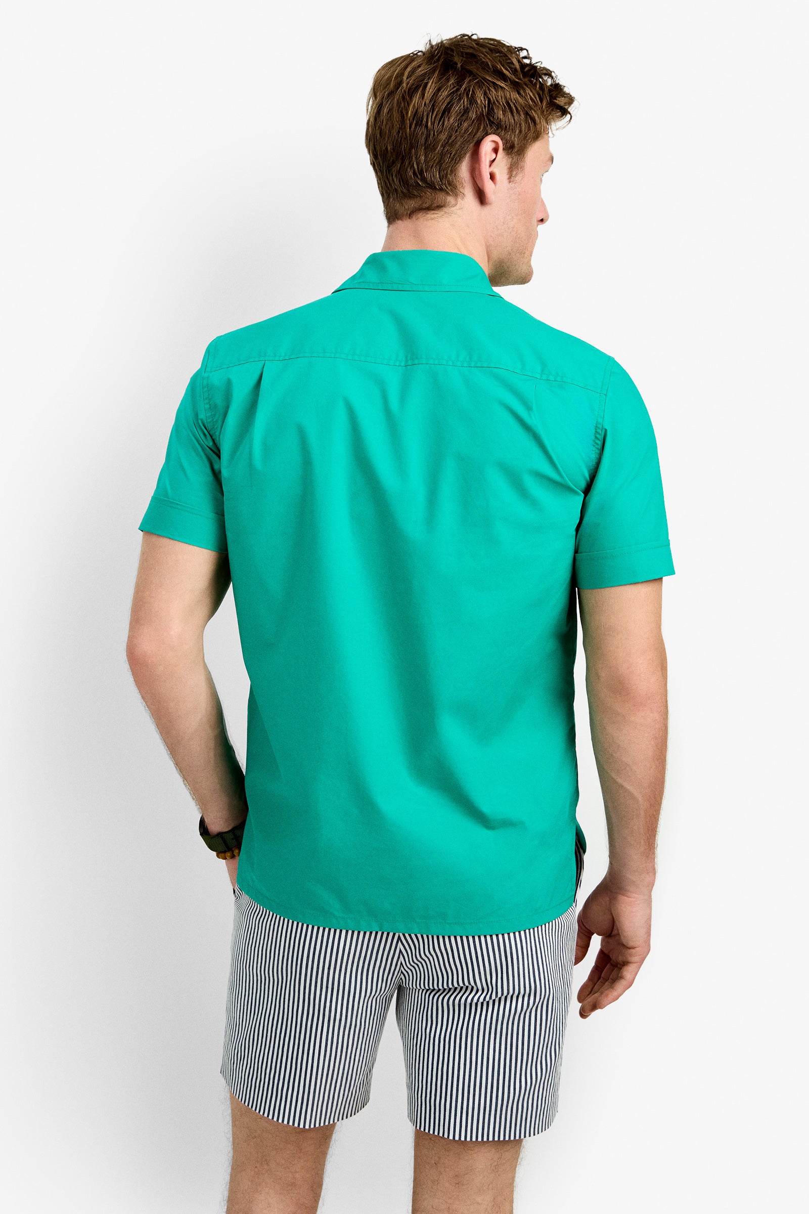 A man faces away against a plain white background, wearing the Will Shirt—a teal short-sleeve button-up—and black-and-white vertically striped shorts.