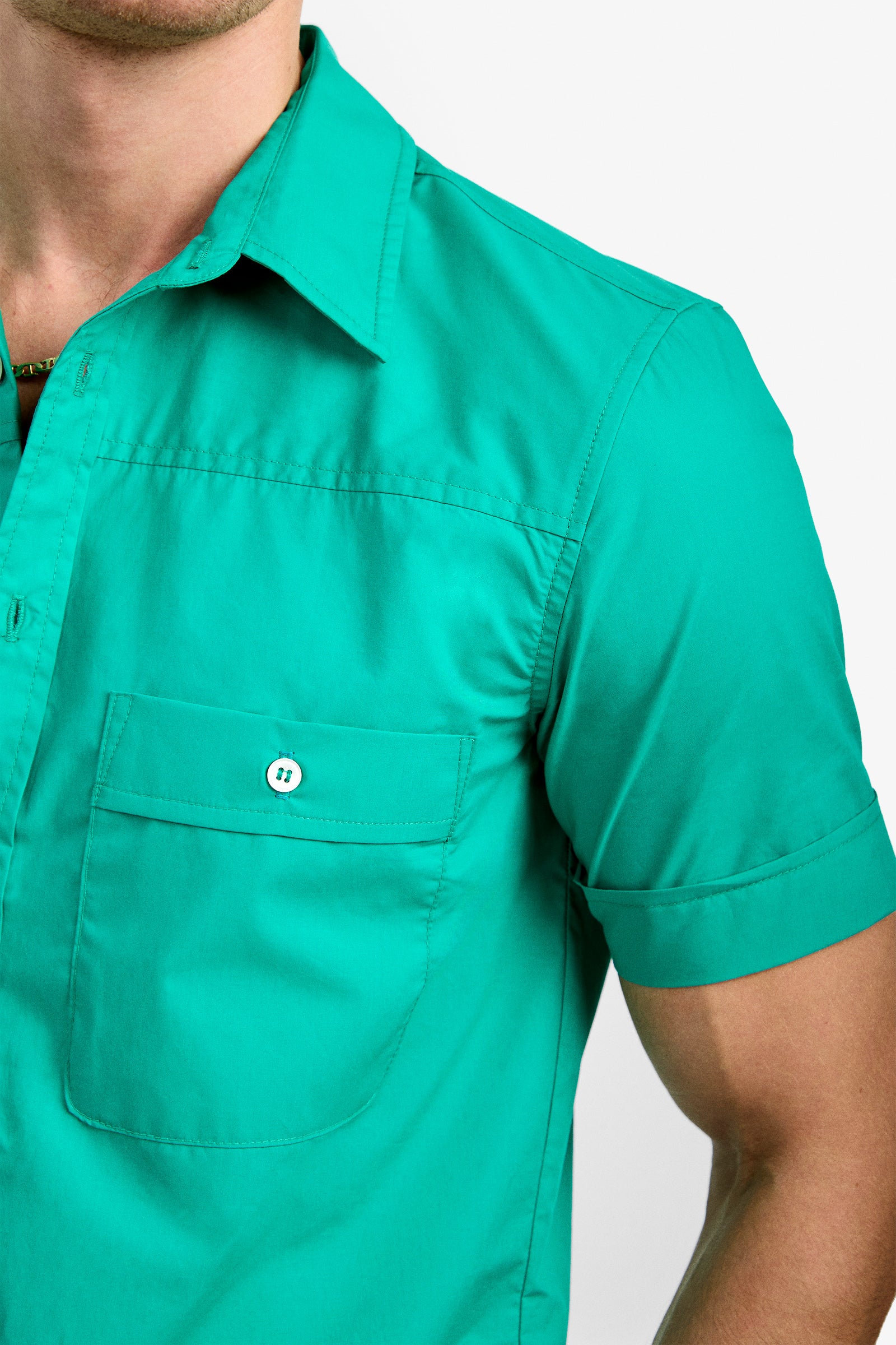 A close-up of a person wearing the Will Shirt, a bright teal short-sleeve button-up with a chest pocket, photographed from chest to shoulder against a plain white background.