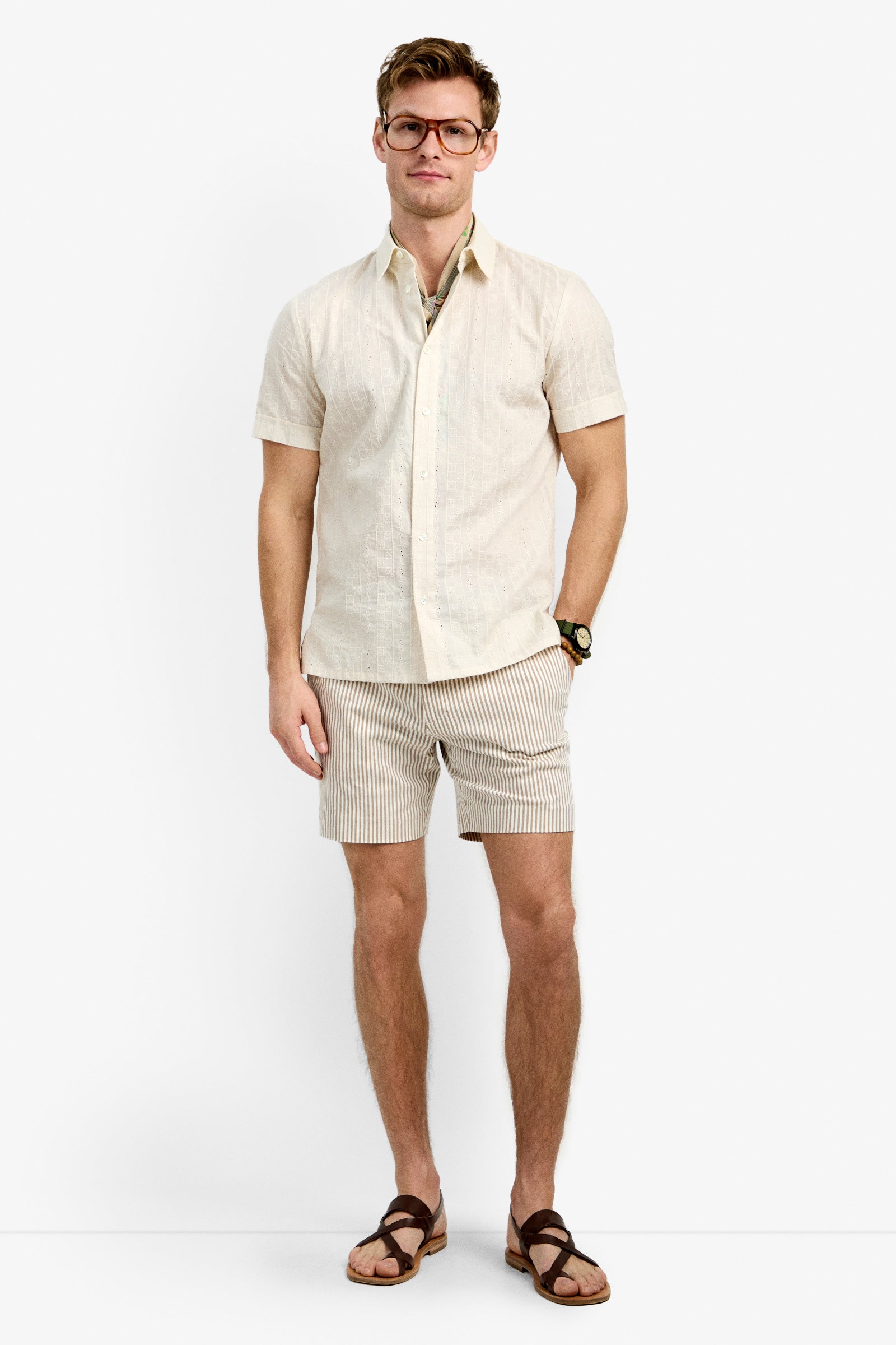 A man wearing glasses, the Branson Shirt (a short-sleeve beige button-up), beige and white striped shorts, brown sandals, and a wristwatch stands against a plain white background, facing the camera with a slight smile.