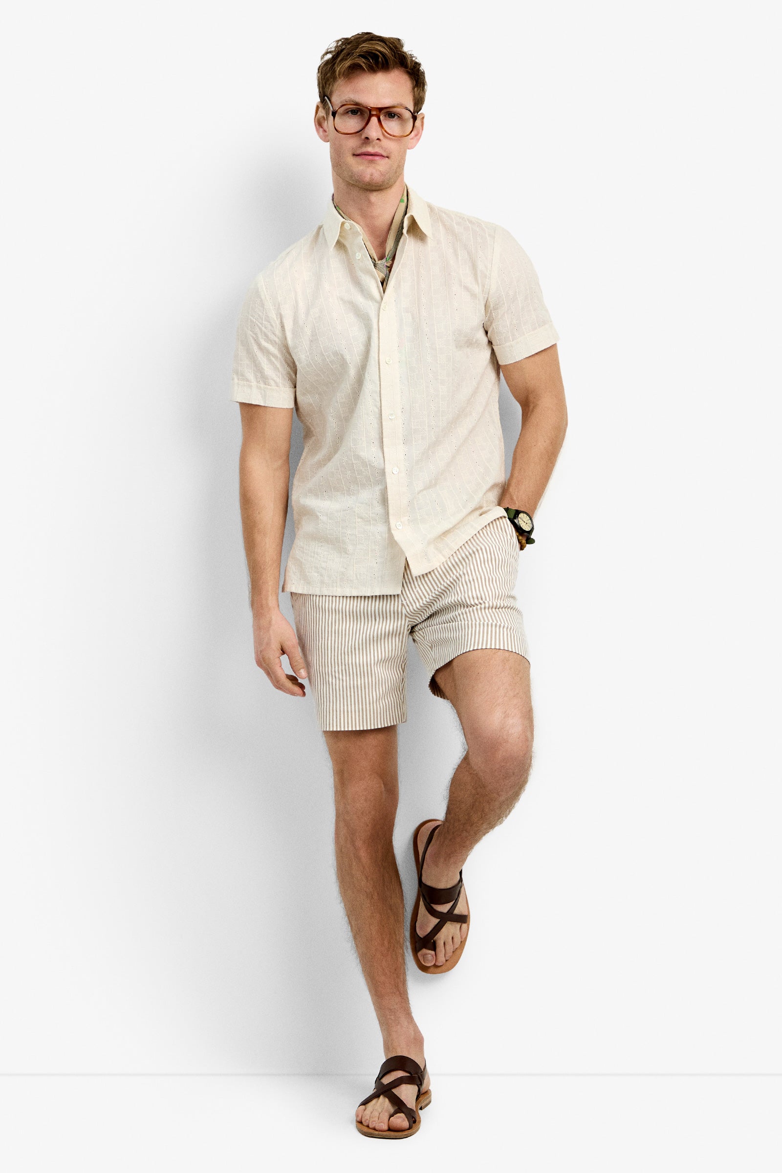 A man stands against a white background wearing the Branson Shirt in Ecru, beige and white striped shorts, brown sandals, glasses, and a wristwatch.
