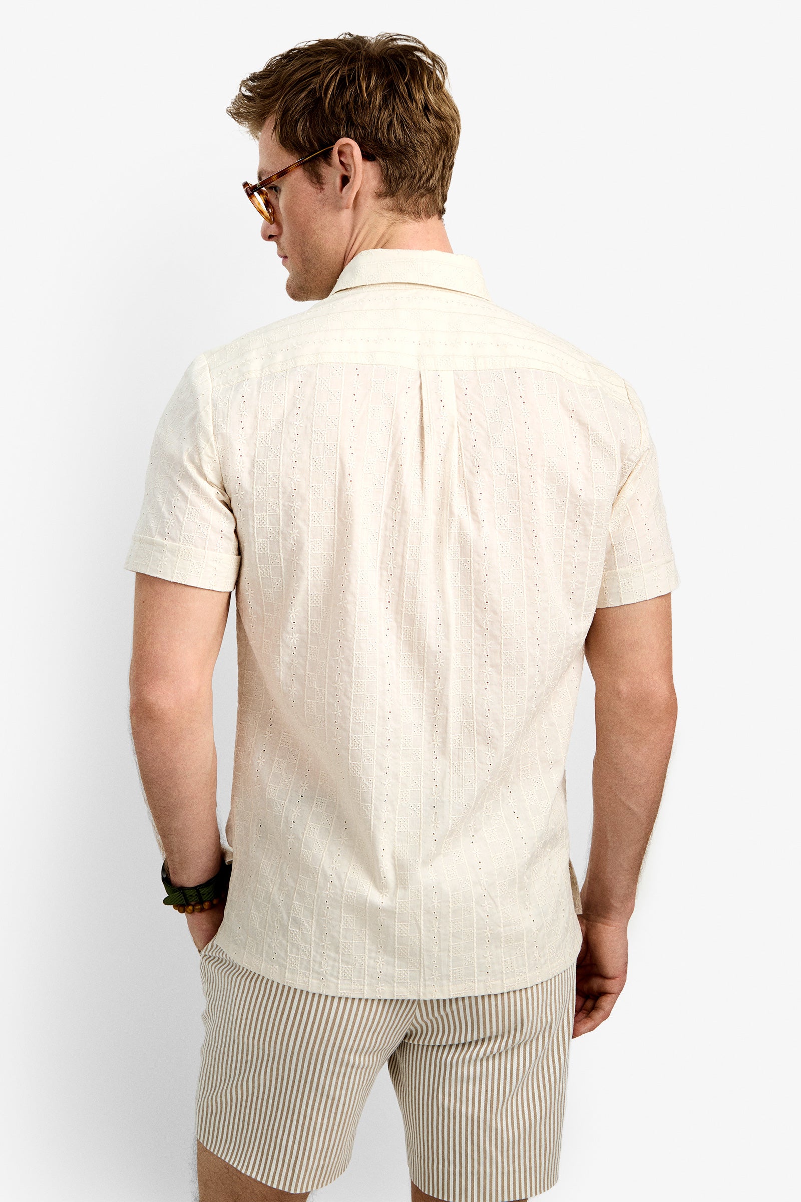 A man with light brown hair wearing glasses and the Branson Shirt, a cream-colored short-sleeve button-up, along with striped beige shorts, stands facing away from the camera against a plain white background.