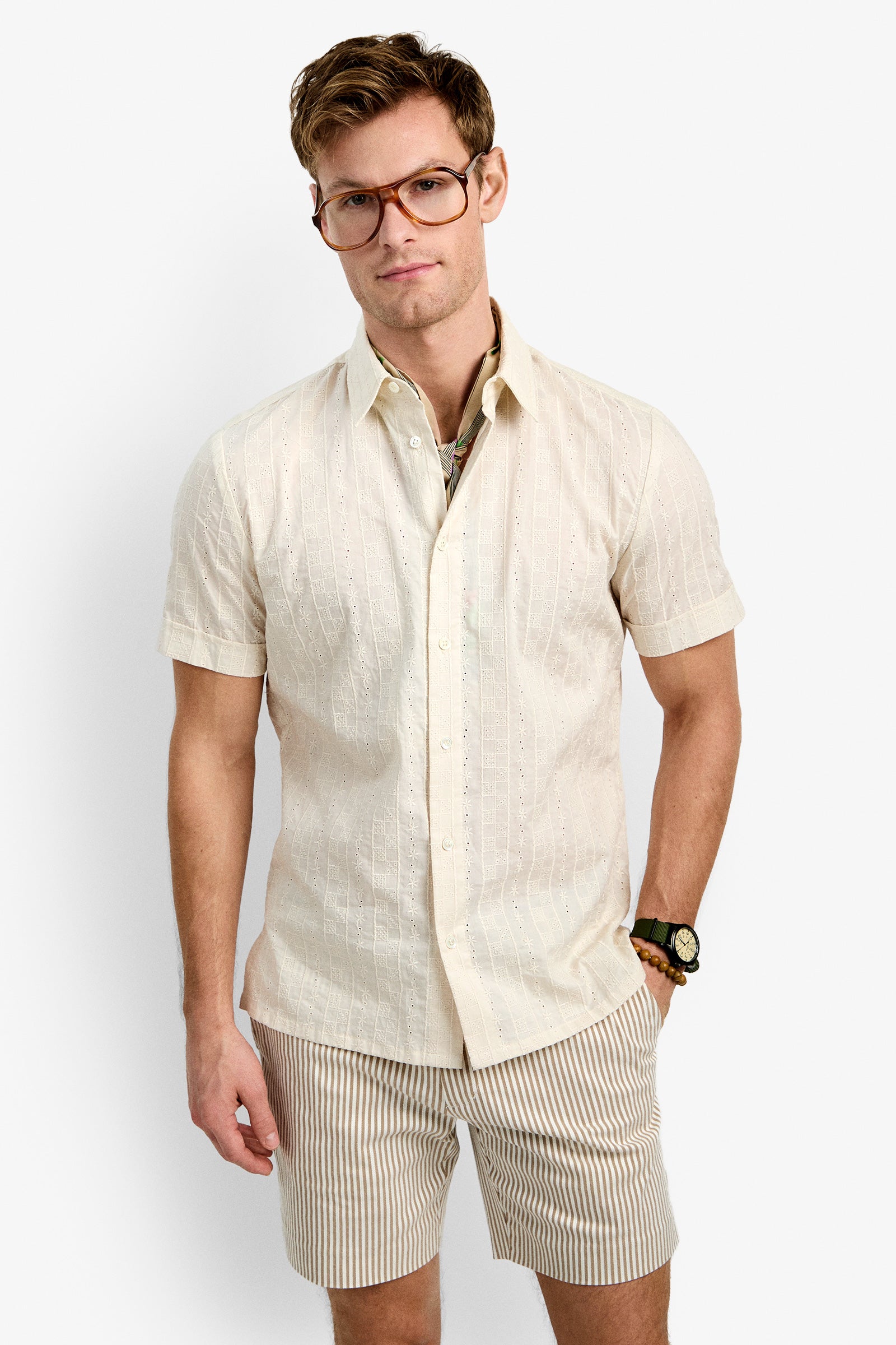 A man wearing the Branson Shirt, a light short-sleeve button-up, stands against a plain white background. He pairs it with light striped shorts and large glasses, looking at the camera with a slight smile.
