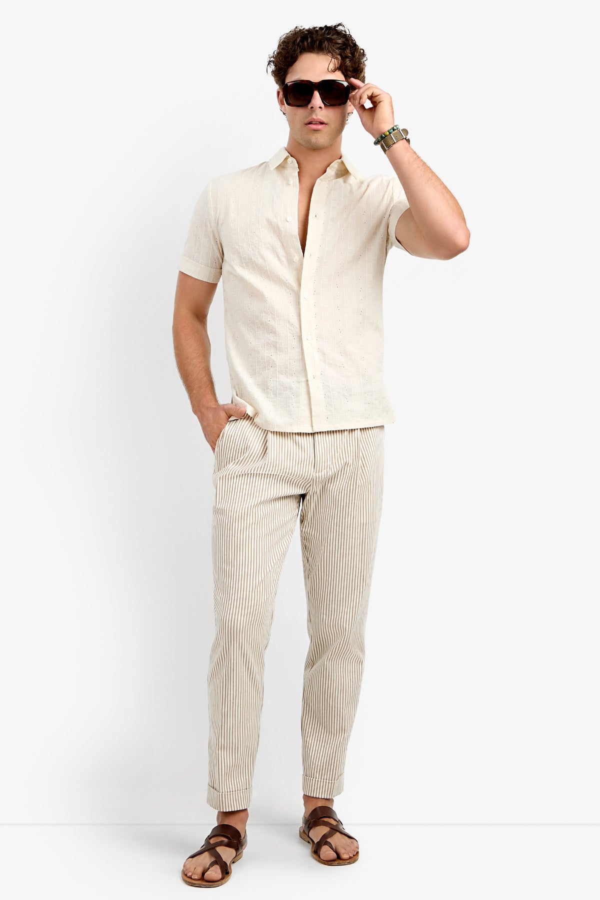 A man wears the Branson Shirt, a short-sleeve beige button-up, paired with tan and white striped pants and brown sandals as he poses with one hand in his pocket against a plain white background.