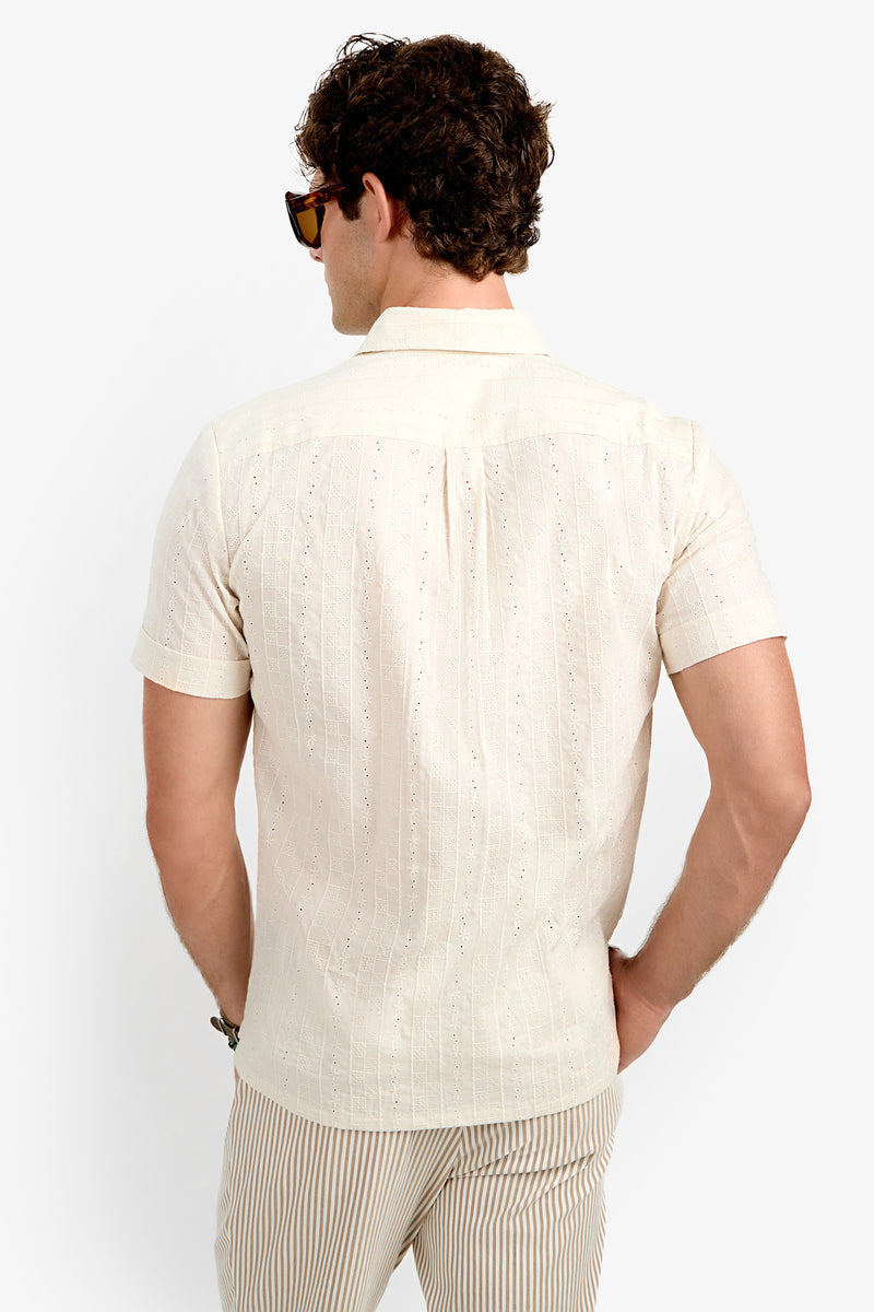 A man with curly hair and sunglasses is seen from behind wearing the Branson Shirt, a light beige short-sleeve shirt with a subtle pattern, paired with beige striped pants against a plain white background.