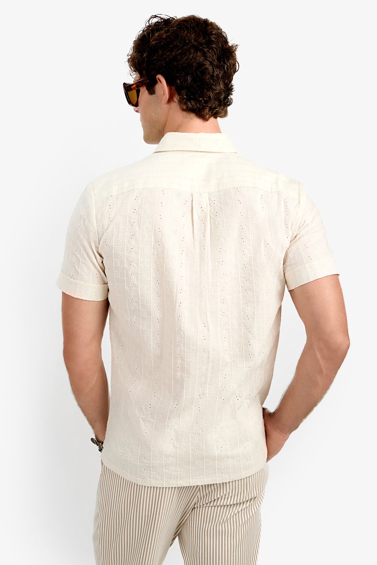 A man with curly hair and sunglasses is seen from behind wearing the Branson Shirt, a light beige short-sleeve shirt with a subtle pattern, paired with beige striped pants against a plain white background.