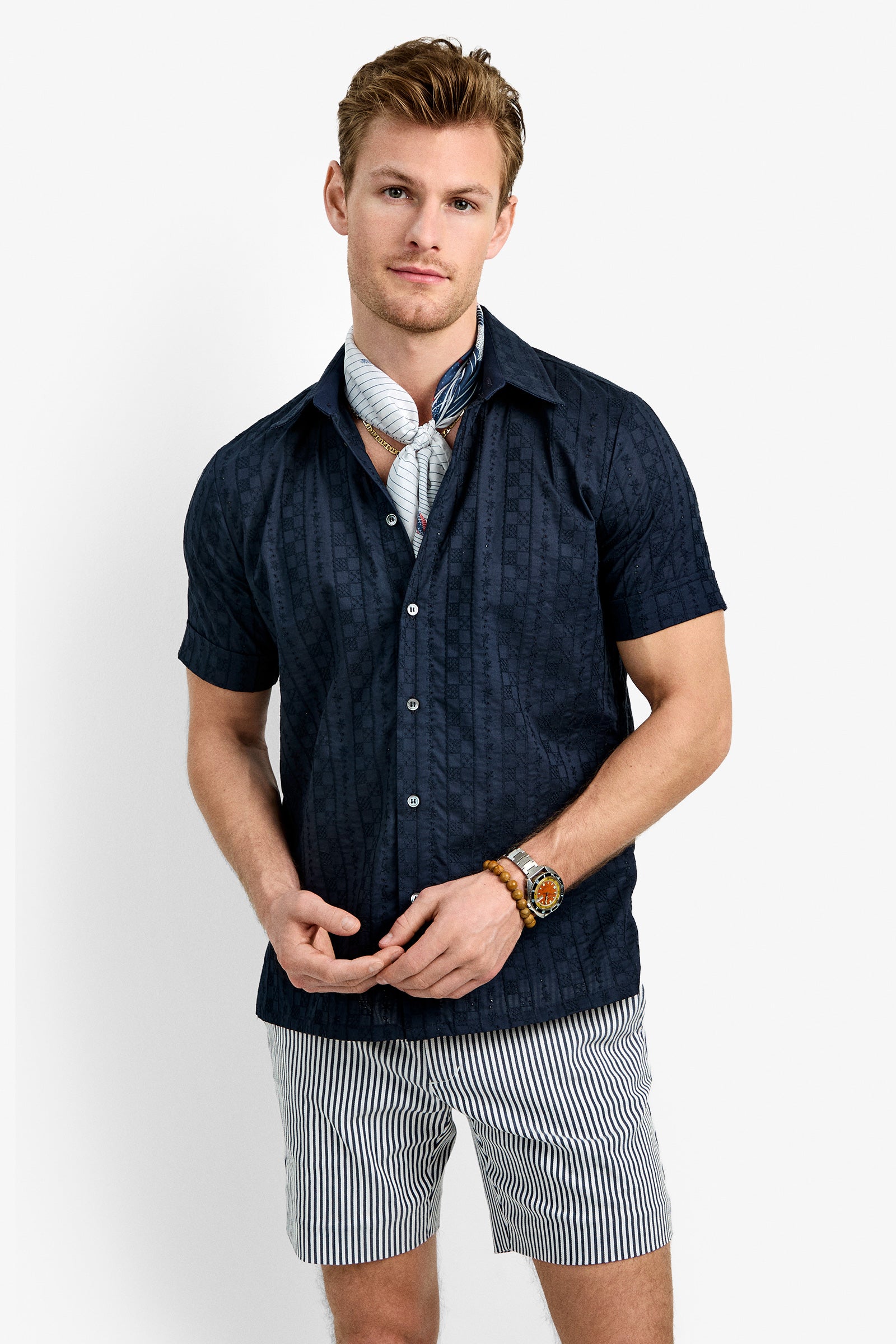 A man wearing the Branson Shirt, a navy short-sleeve button-up, paired with light striped shorts, a light scarf, and a watch with bracelets stands against a plain white background.