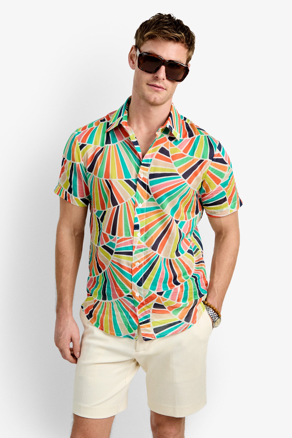 A man wearing the Slim Jim Shirt—a colorful short-sleeve button-up with a geometric pattern—large dark sunglasses, and light beige shorts stands against a plain white background.
