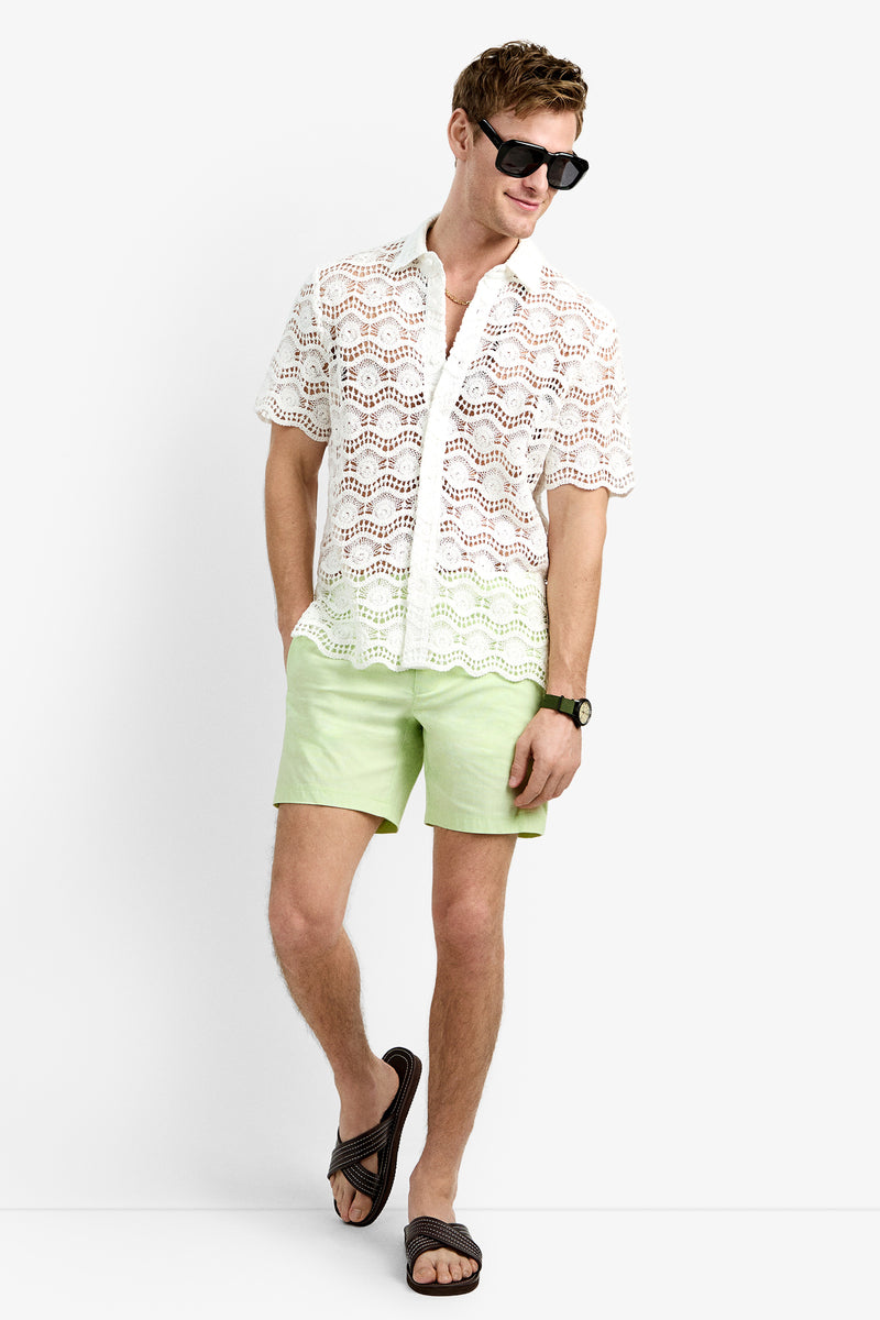 A man wearing black sunglasses, a WHITEWASH Long Beach Shirt, light green shorts, a black wristwatch, and black slide sandals stands against a plain white background.