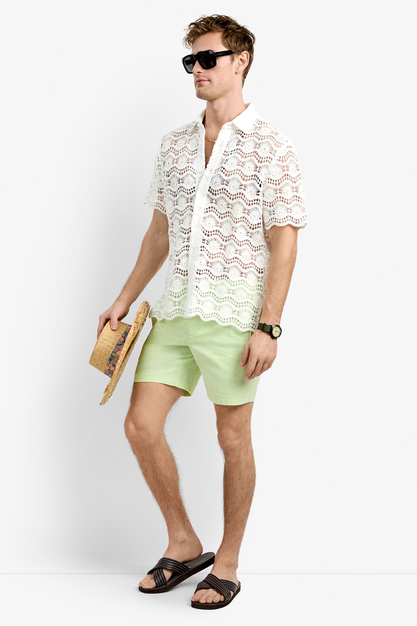 A man with short light hair poses against a plain white background wearing sunglasses, light green shorts, black sandals, a watch, and the Long Beach Shirt. He stands holding a straw hat.