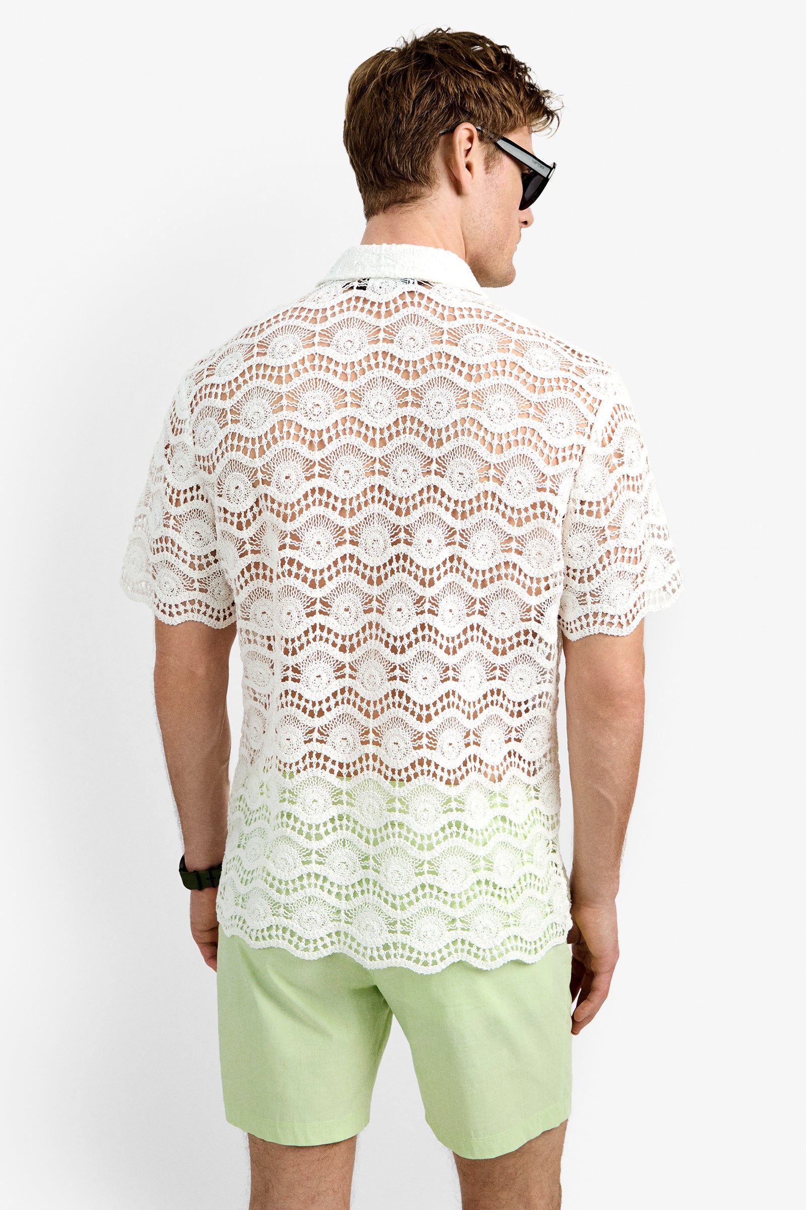 A man faces away from the camera against a plain white background, wearing black sunglasses, light green shorts, and the Long Beach Shirt, a white short-sleeve crochet top.