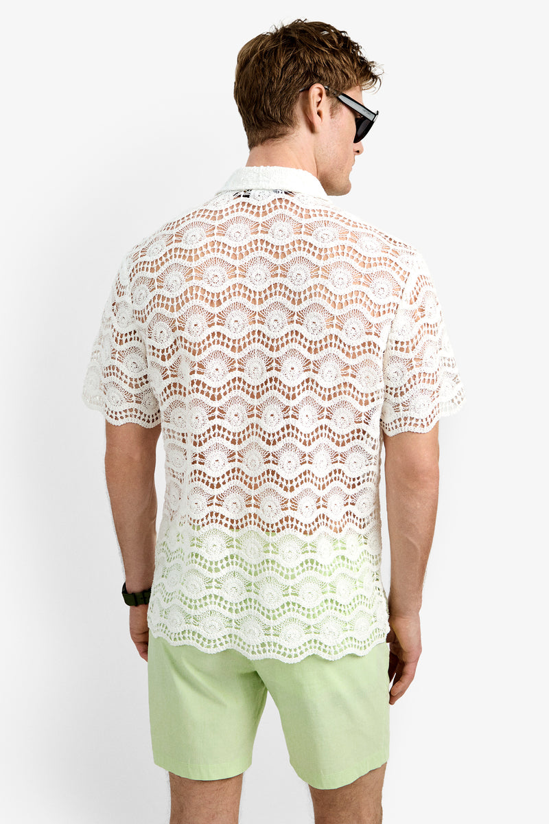 A man faces away from the camera against a plain white background, wearing black sunglasses, light green shorts, and the Long Beach Shirt, a white short-sleeve crochet top.