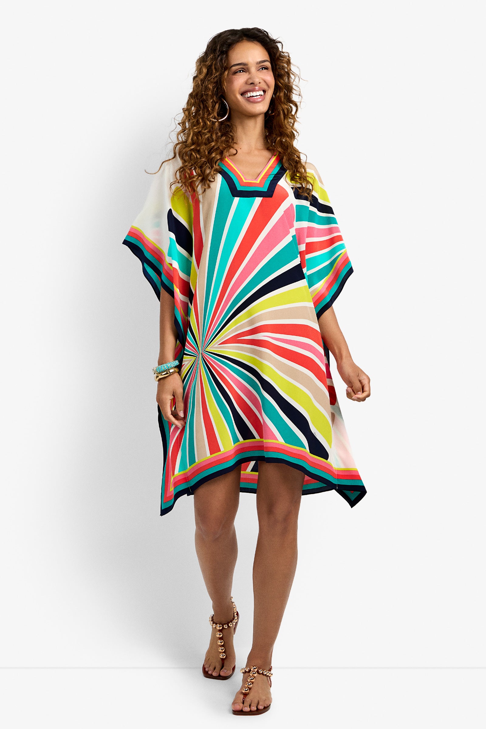 A woman with curly hair smiles as she walks in the Theodora Dress—a loose, short-sleeved dress with bold, colorful radiating stripes—paired with sandals and bracelets against a plain white background.