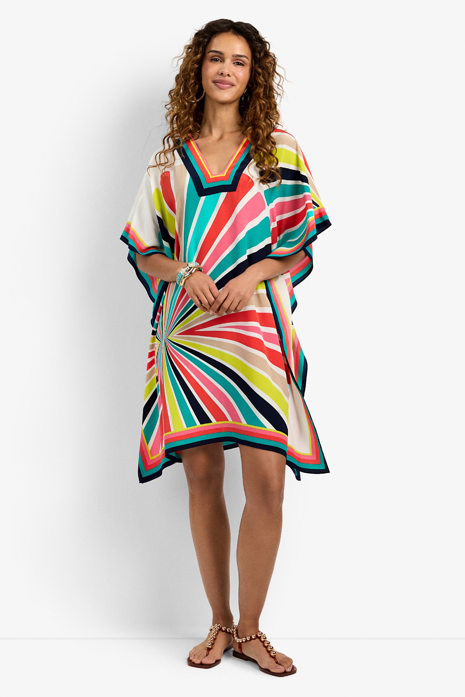 A woman wears the Theodora Dress in MULTI, a colorful, loose-fitting style with bold radiating stripes, standing and smiling against a plain white background.