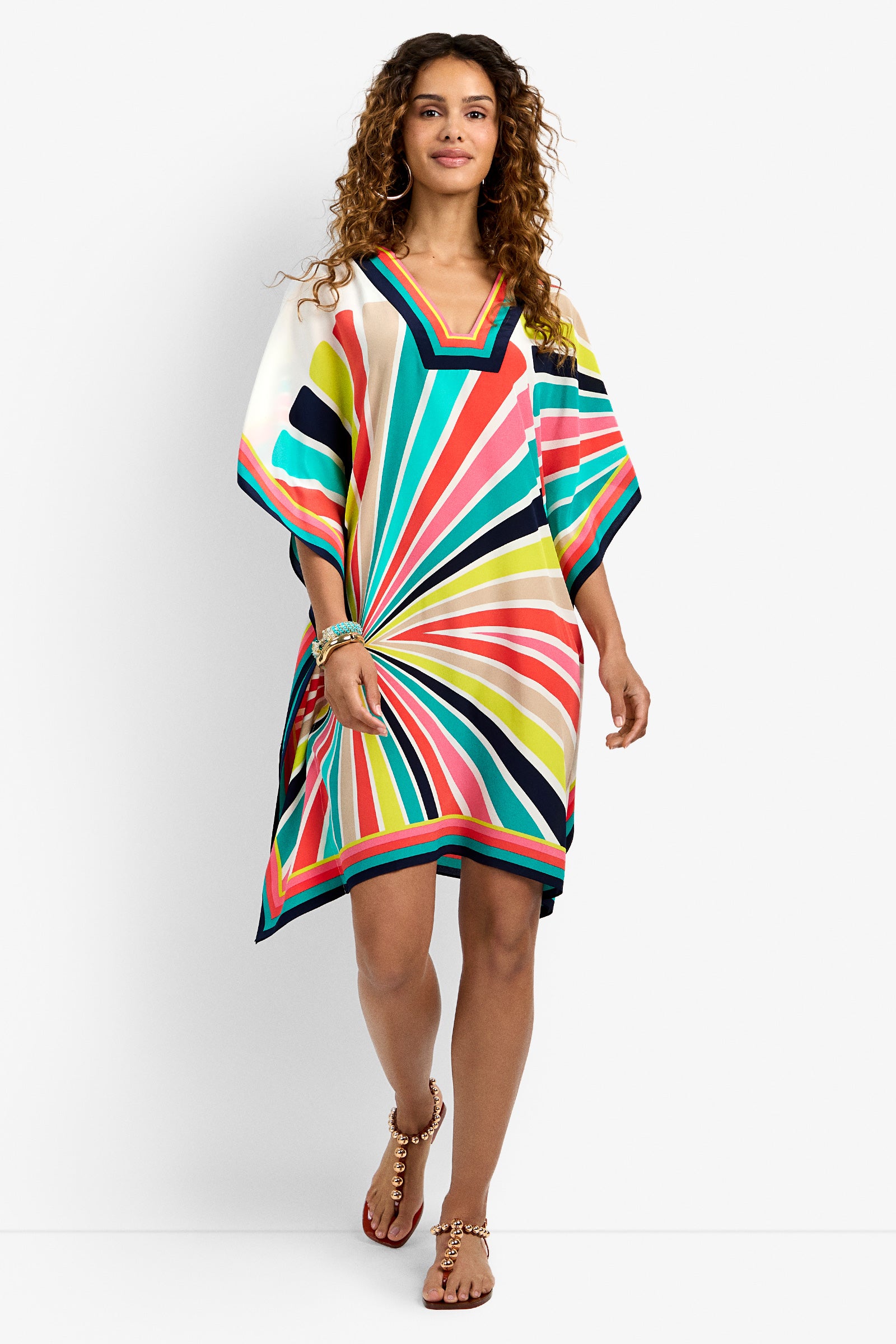 A woman with curly hair smiles and walks confidently against a white background, wearing Theodora Dress—a colorful, striped, knee-length kaftan with short sleeves and a V-neck—paired with sandals.